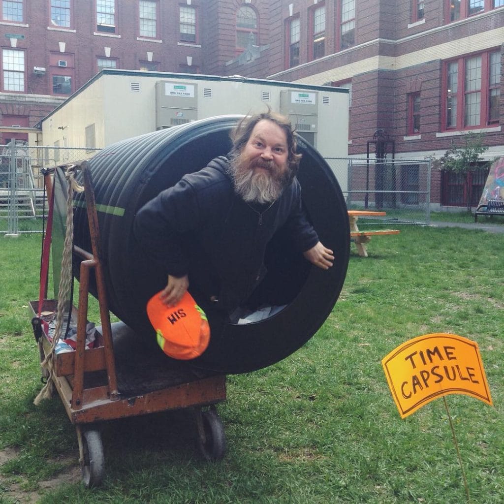 A bearded man playfully emerges from a large cylindrical tube labeled as a Time Capsule. He holds an orange cap marked HIS. The scene is outdoors on grass, near a brick building with picnic tables in view. An orange sign reads TIME CAPSULE.