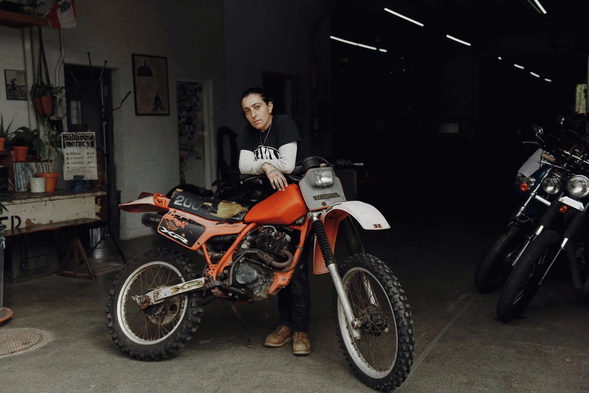 Person leaning on an orange dirt bike inside a dimly lit garage, exuding balance and cool confidence. Clad in casual attire, they are surrounded by several other motorcycles. Potted plants and framed pictures artfully decorate the walls, adding a touch of warmth to the mechanical haven.