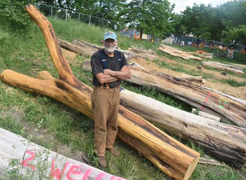 An older man with a beard and cap stands confidently in a grassy area of Danehy Park, surrounded by large, cut logs. He wears a black shirt, suspenders, and brown pants. A fence and the lush trees of Cambridge provide a serene backdrop.