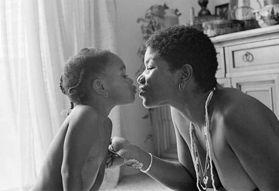 A black and white image captures a tender moment between a young child and an adult woman, likely a mother, sharing a kiss in a warmly lit room. Embracing themes from Designing Motherhood, the scenes background of cabinets and soft curtains adds to its material culture essence.
