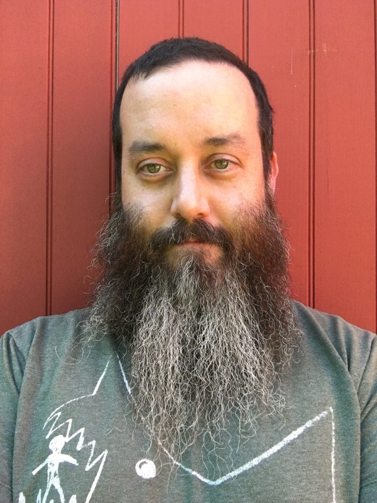 A person with a long, full beard and short hair stands against a red wooden background. They are wearing a gray shirt with a white graphic design and have a calm expression.