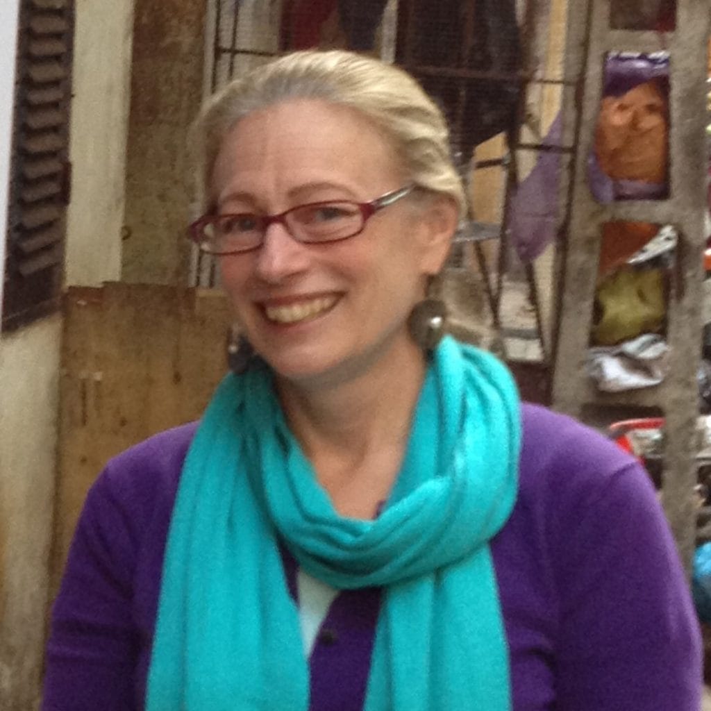 A person with light hair tied back is smiling, wearing glasses, a purple top, and a turquoise scarf. They are standing outdoors in an urban setting with a blurred background of buildings and hanging laundry.
