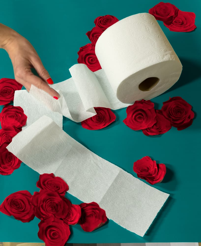 A hand with red-painted nails unrolls a strip of toilet paper on a teal surface, surrounded by scattered red roses, reflecting the creativity sparked during COVID Projects.