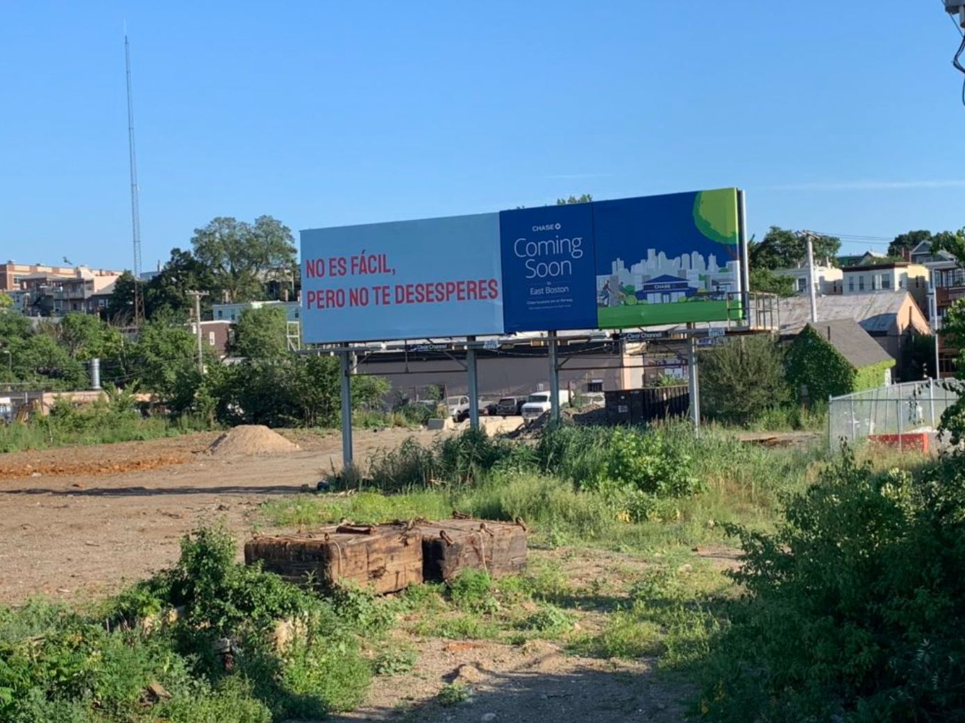 A billboard crafted by Gabriel Sosa stands in an open area, saying No es fácil, pero no te desesperes beside a cityscape illustration and Coming Soon, East Boston. This message of hope is surrounded by trees, buildings, and patches of dirt.