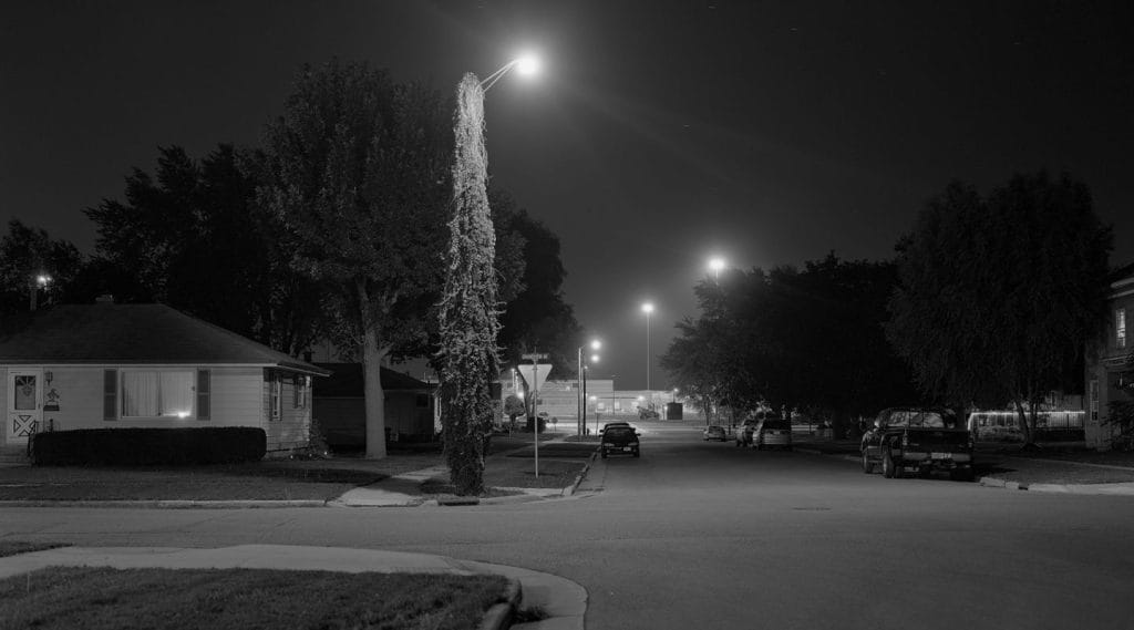 A quiet residential street at night in grayscale. A single streetlight illuminates the area, reminiscent of the somber aura surrounding a correctional facility. A few parked cars and houses line the road, while distant lights dot the clear sky in the background.