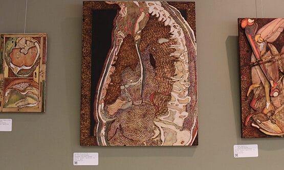 Three mixed media artworks by Brett Poza adorn the wall, featuring abstract anatomical designs in earthy tones. Small labels with QR codes accompany each piece. The central artwork is larger, flanked by two smaller, complementary pieces showcasing his pyrography technique.