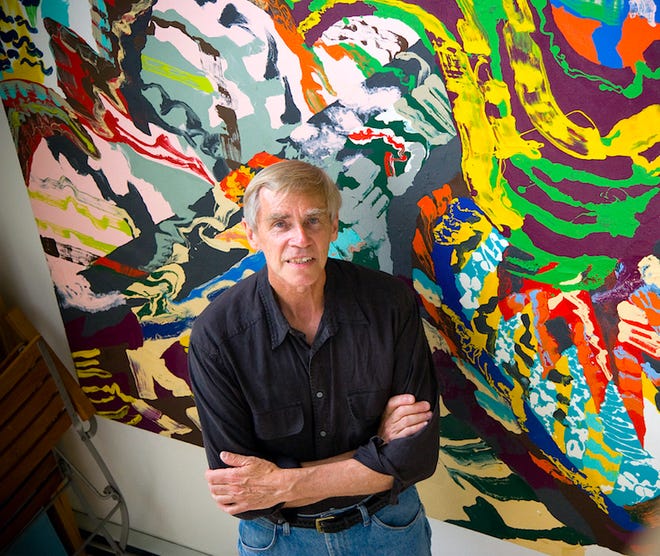 A person in a black shirt and blue jeans stands with arms crossed in front of a colorful abstract painting by Southcoast artist Roger Kizik. The artwork, showcased at Art Beat, features a chaotic swirl of vibrant colors like yellow, green, red, and blue on a large canvas.