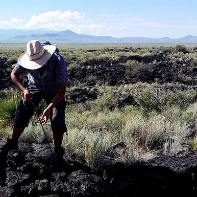 A person wearing a wide-brimmed hat examines rocky terrain with a tool in hand. The landscape features sparse vegetation and distant mountains under a clear blue sky.