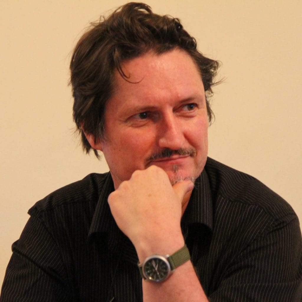 A man with wavy brown hair and a short beard is resting his chin on his hand. He is wearing a black shirt and a wristwatch with a green strap. The background is a plain, light-colored wall.