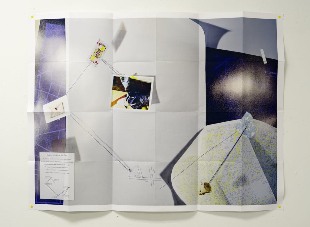 A large sheet of paper is pinned on a wall, displaying a makeshift map with connected images and notes. Strings link various points, with fingerprints highlighted in the east section. The layout resembles a plan or investigation board.