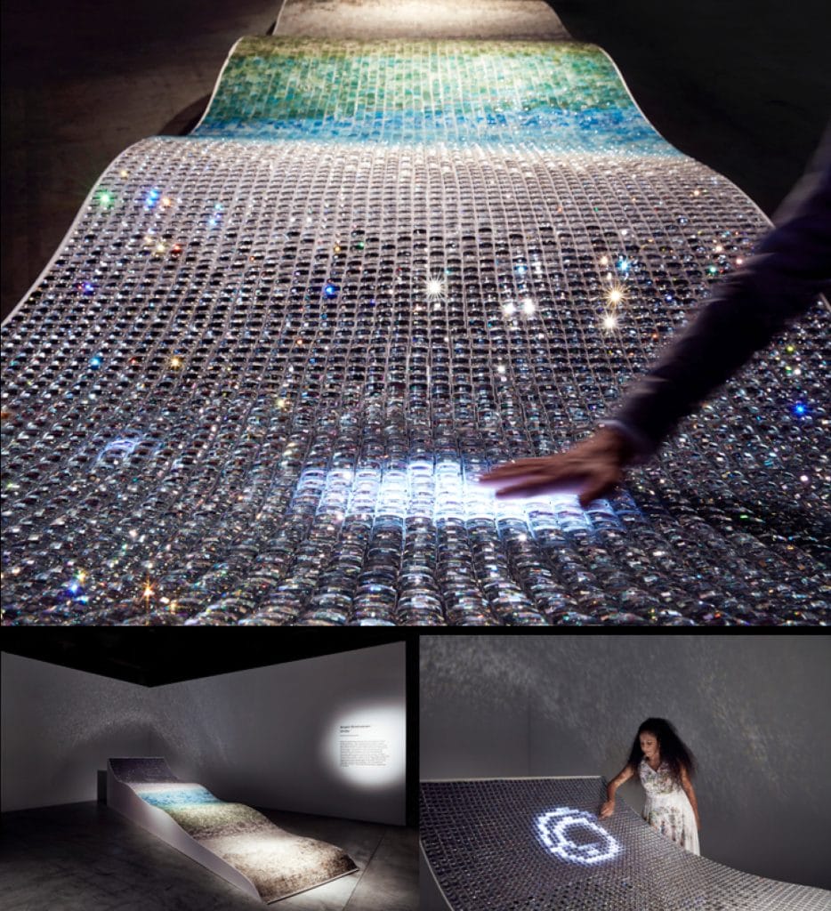 A glowing installation with a textured, undulating surface covered in transparent spheres. A person interacts with the piece, which emits light patterns and colors. The display is set in a dimly lit gallery space, creating an immersive atmosphere.