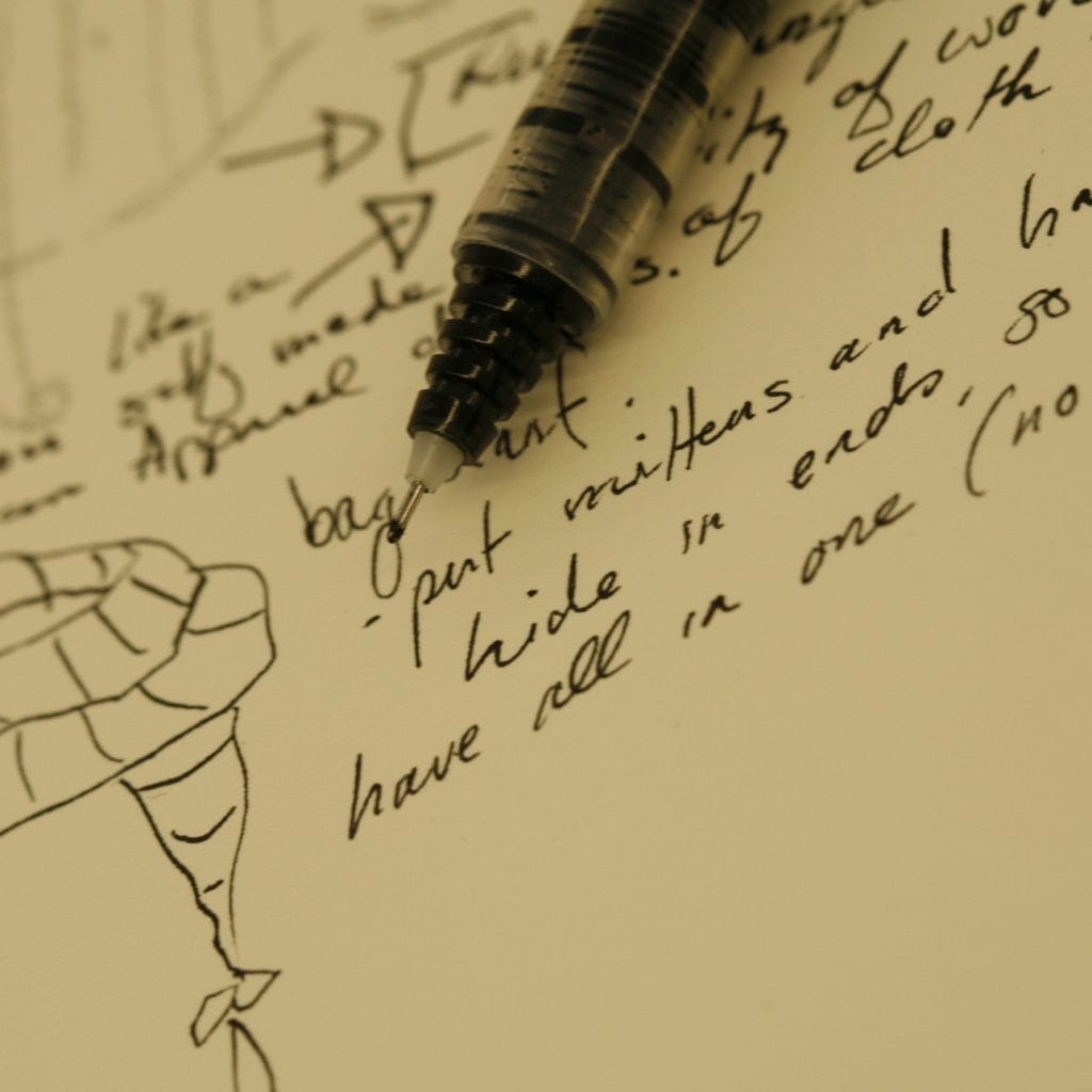 Close-up of a pen resting on a piece of paper with handwritten notes and a drawing of a striped object, possibly a scarf or fabric. The notes include phrases like put mittens and hide in ends and have all in one.