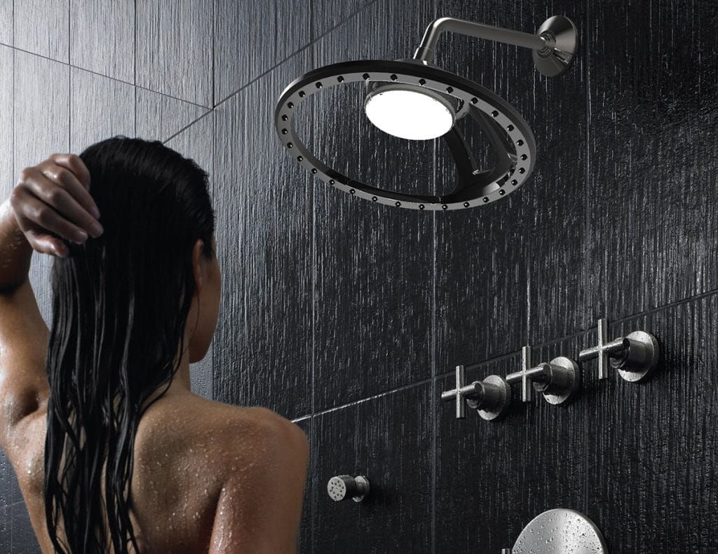 A person with wet hair stands under a modern circular showerhead, enhanced by Sunshower Light Therapy, in a dark-tiled bathroom. The water cascades down as the soothing LED light glows, and three metal knobs gleam on the wall.