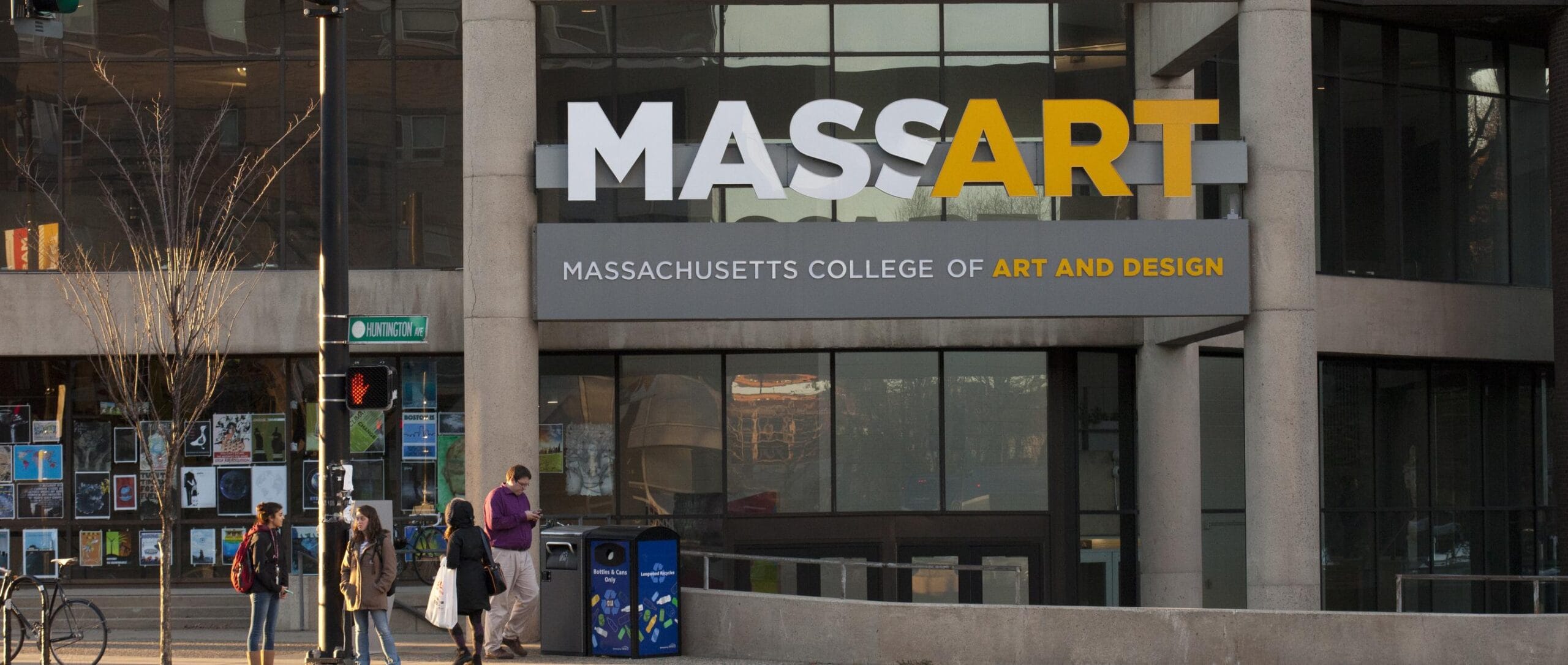 The image captures the entrance of the Massachusetts College of Art and Design. A prominent MASSART sign stands tall as people stroll along the sidewalk. Posters in the windows create a vibrant playbook of creativity and innovation, embodying the spirit of Massachusetts artistry.