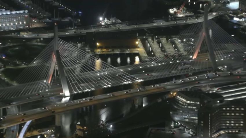 An inaugural aerial view of a suspension bridge illuminated at night, stretching over a dark waterway. The bridges cables fan out from its central towers, with city lights reflecting below. This Boston site offers a serene tribute to the citys resilience during the COVID-19 memorial events.