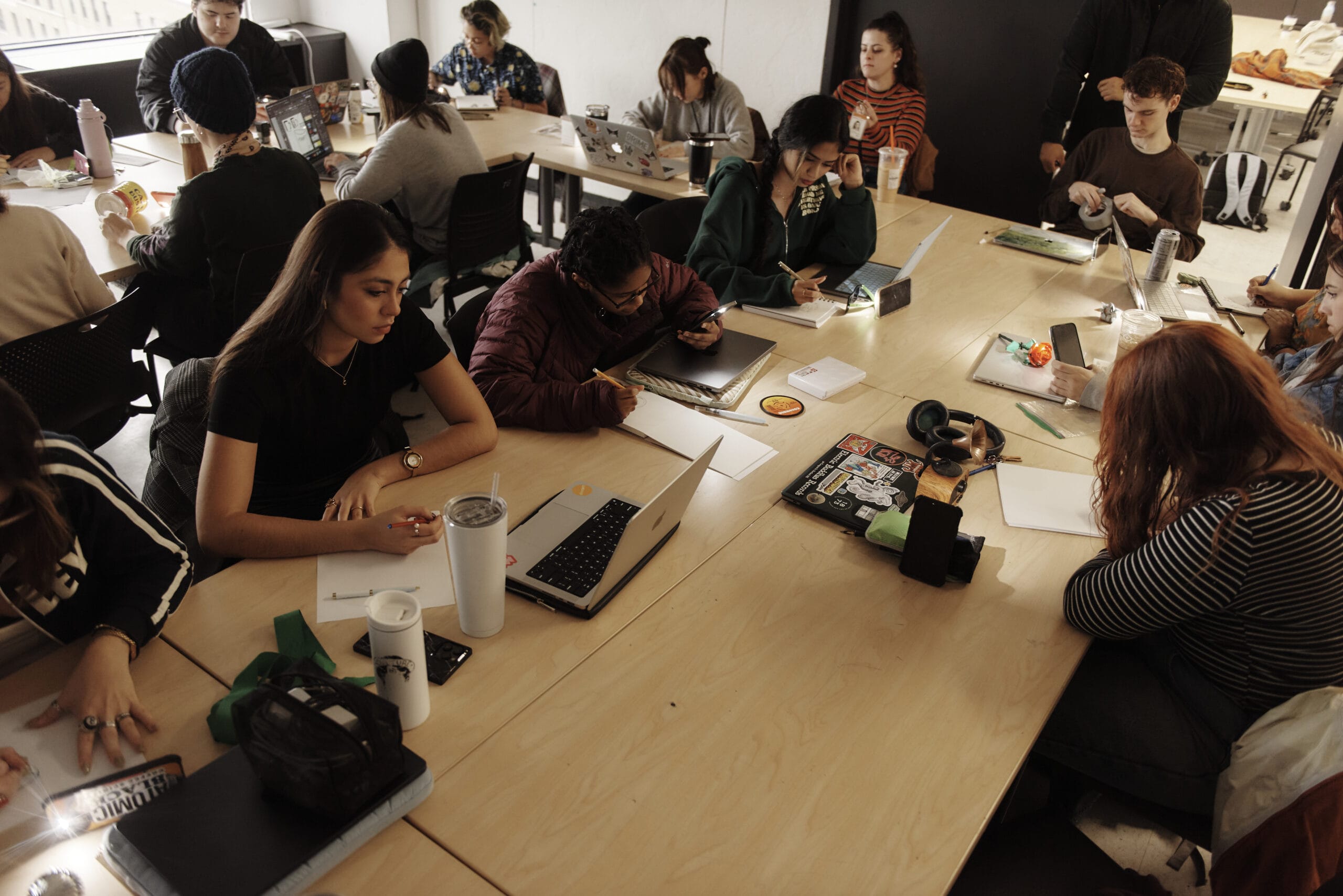 Students sit at an L shaped table looking at their laptops and working in a classroom.