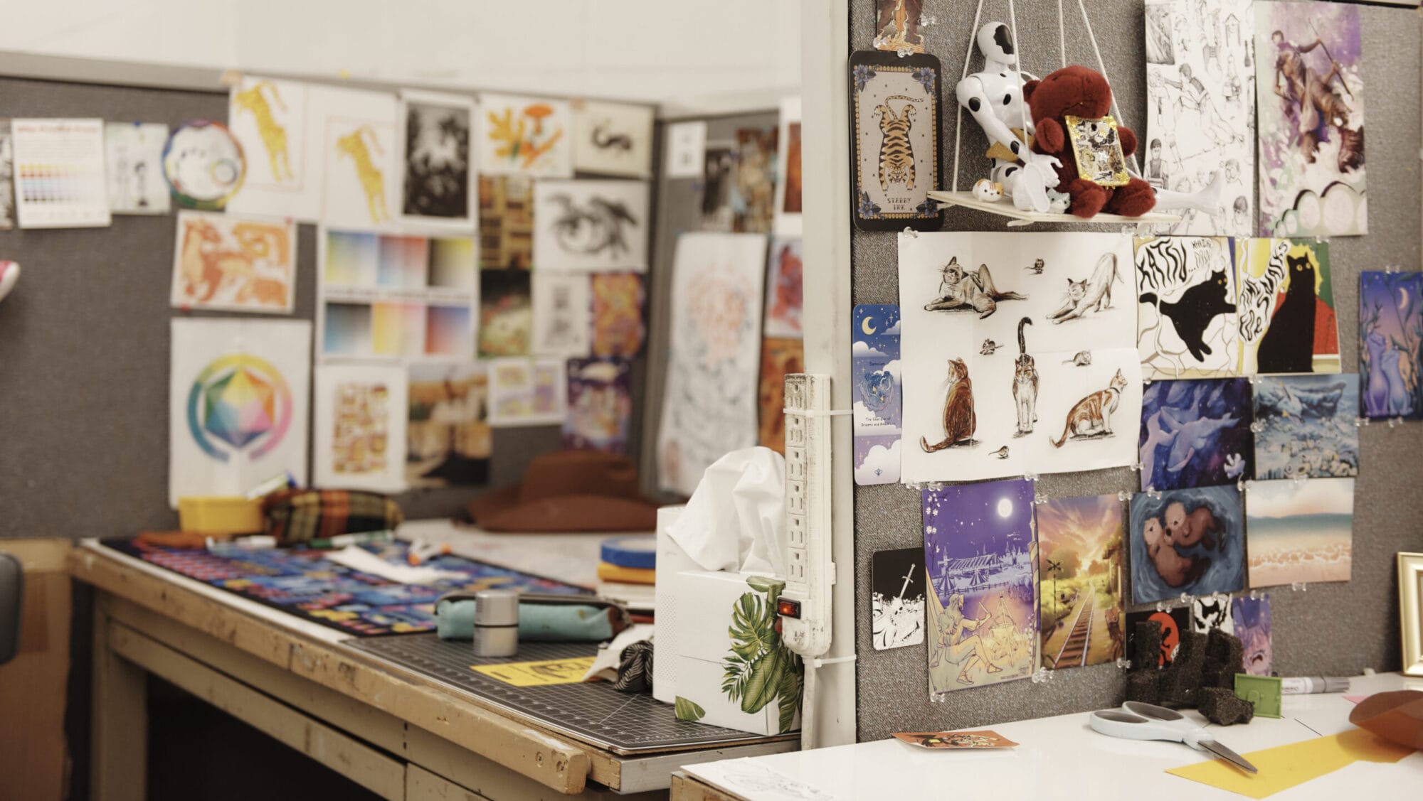 An art studio desk with colorful supplies, posters, and a BFA Illustration certificate pinned to partitions. The workspace is cluttered with paint, brushes, and art books, surrounded by vibrant and diverse artwork, including animals and abstract designs.