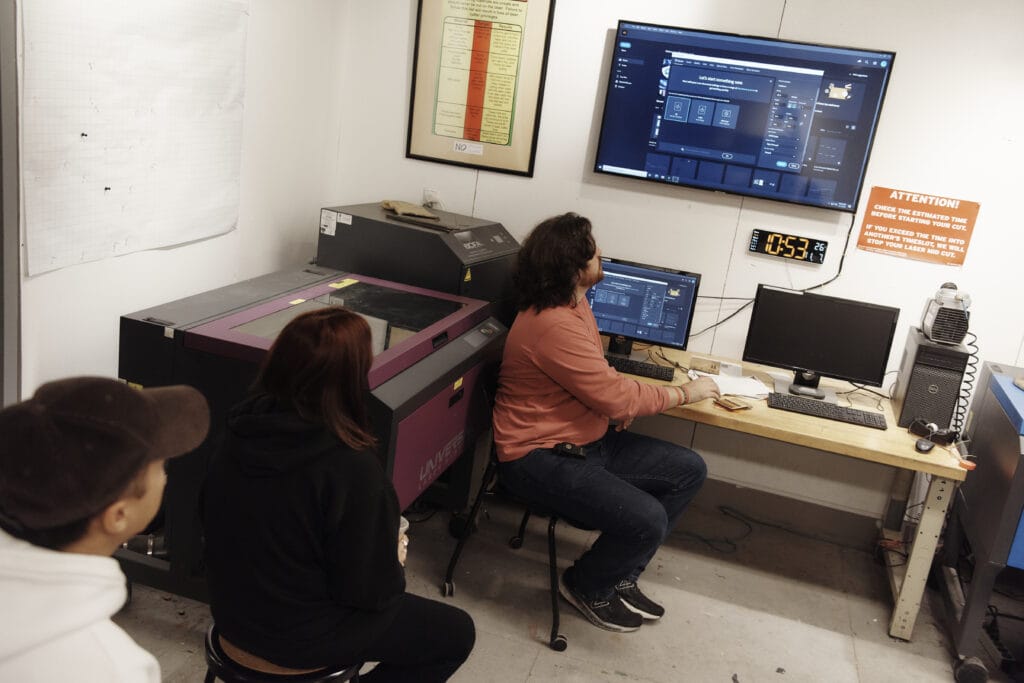 Three people are in a studio filled with electronic equipment. One person is seated, operating a computer connected to a laser machine, while two others watch. A large monitor displays software. A framed chart and a sign about resources adorn the walls.