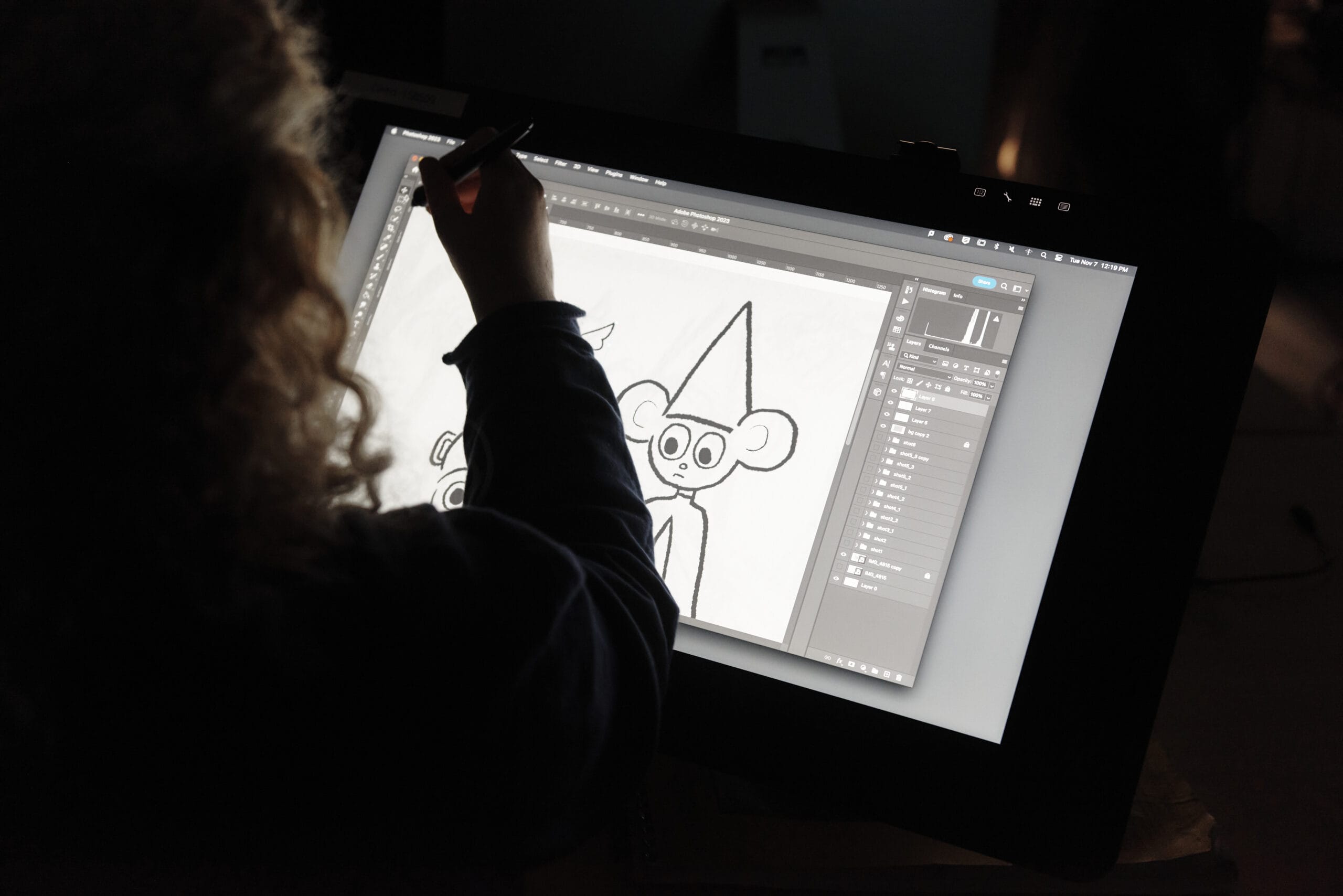 A person with curly hair sketches a cartoonish animal character wearing a pointy hat on a digital drawing tablet. The screen displays drawing tools and layers in an animation studio software interface, set against a dimly lit background.