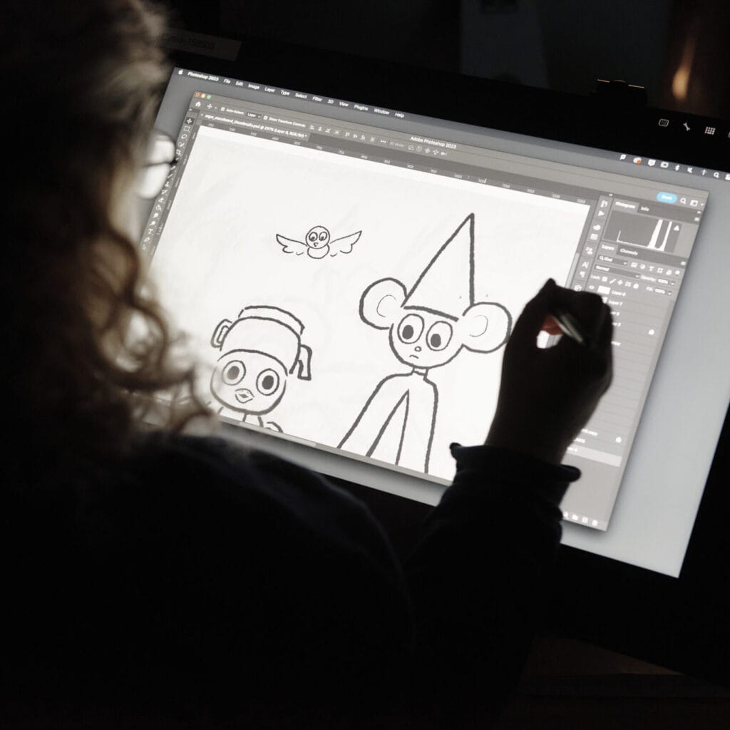 A person uses a stylus on a digital tablet to draw cartoon characters. The screen displays a sketch of a monkey with a tall hat and a dog with a beanie, along with a flying creature above them.