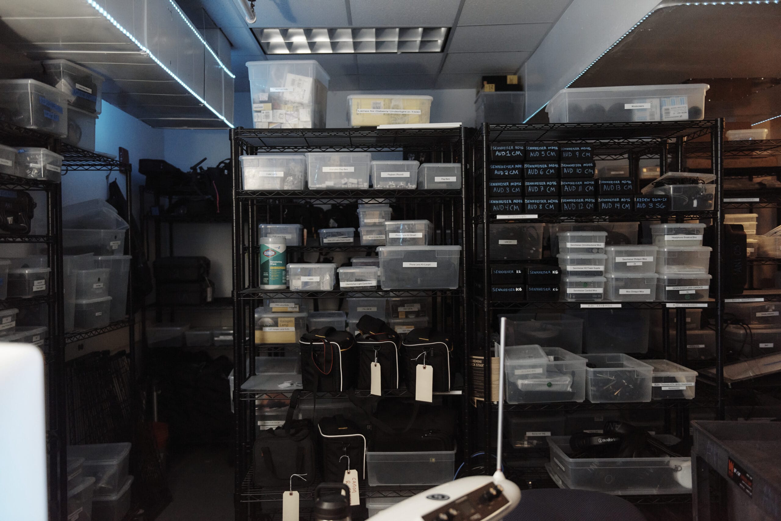 The dimly lit storage room, likely part of the animation studios, features metal shelves packed with labeled plastic bins of various equipment. Bags hang alongside them, and a security camera sits on a desk in the foreground, silently keeping watch over this creative space.