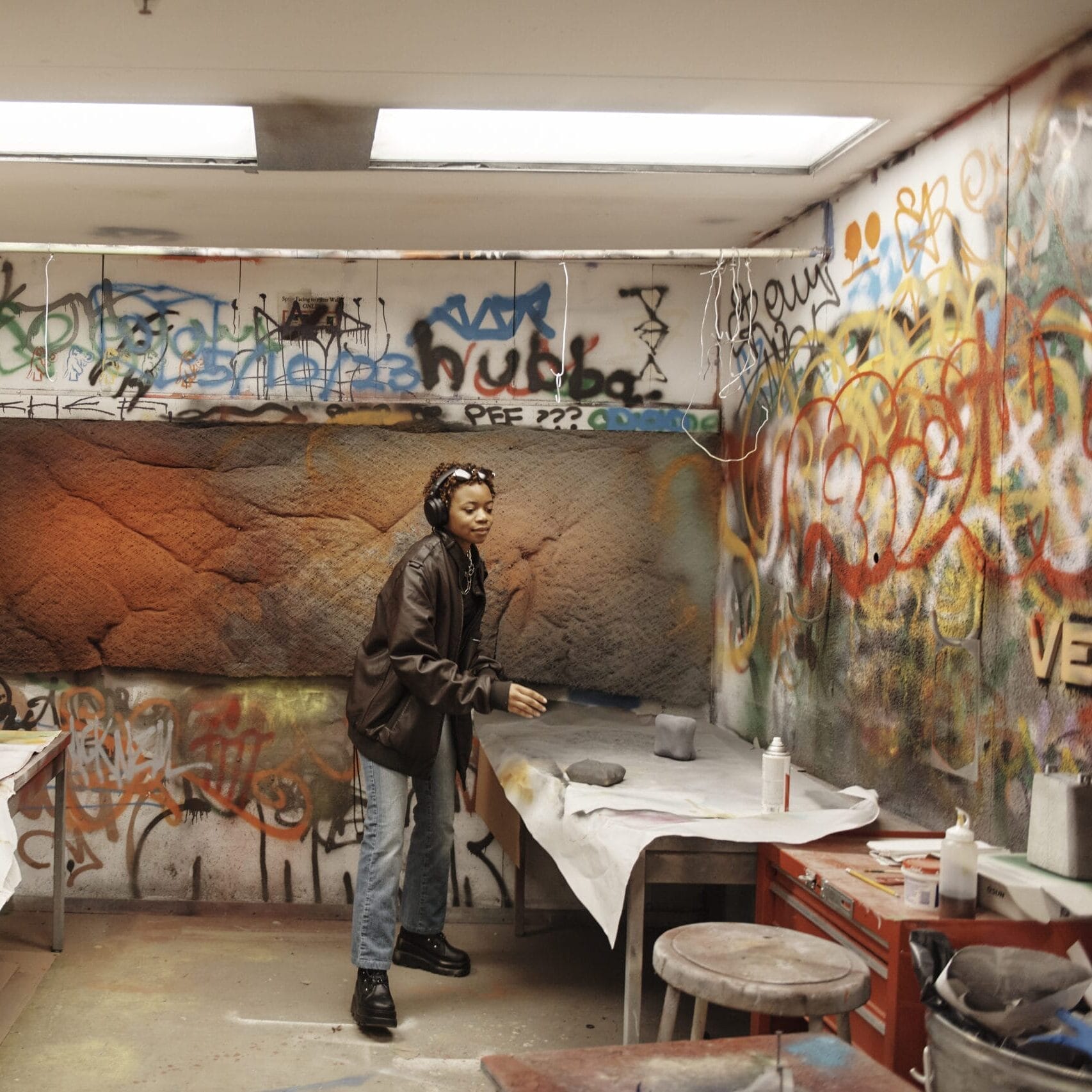 A person wearing a brown jacket and beanie is in a graffiti-covered room, reminiscent of edgy design studios, bending over a table with papers and spray paint cans. Shelving and stools are visible amidst the industrial design vibe, enhanced by fluorescent lights overhead.