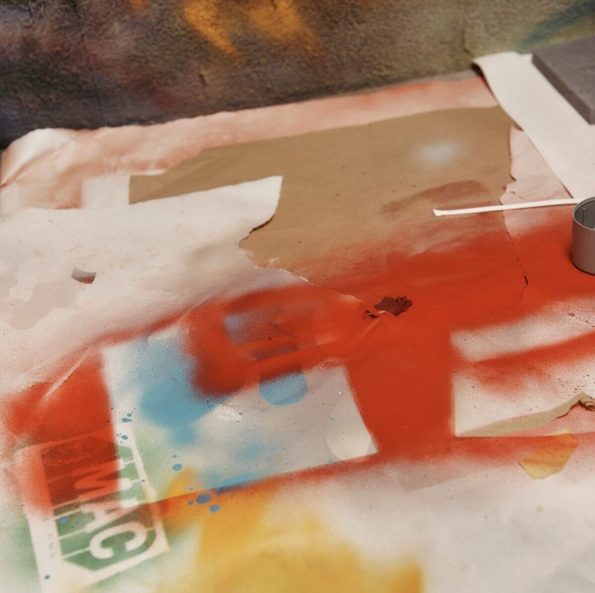 A table in an industrial design studio is covered with scattered paper, spray paint in vibrant colors, and a small metal cylinder. Red, blue, and yellow paint splatters decorate the paper, along with torn edges and smudges. A cotton swab lies nearby.