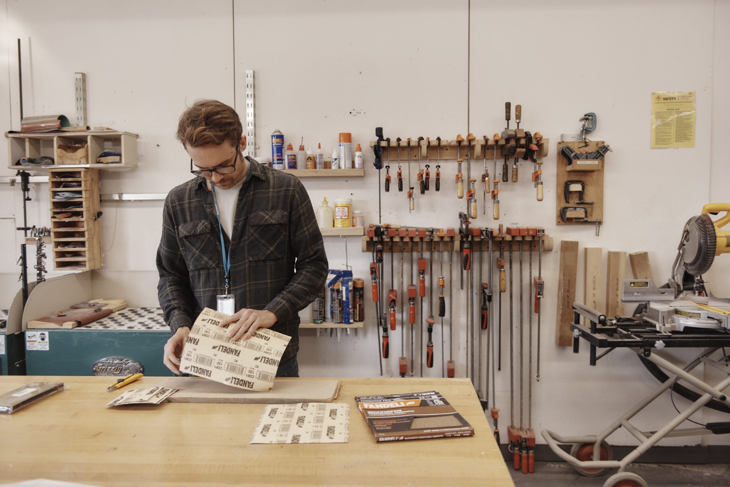 A person in a workshop stands at a table organizing sheets of sandpaper. The background evokes an industrial design vibe with tools like clamps and a saw neatly arranged on the wall. Shelves hold various supplies, reminiscent of design studios, creating a well-equipped workspace.