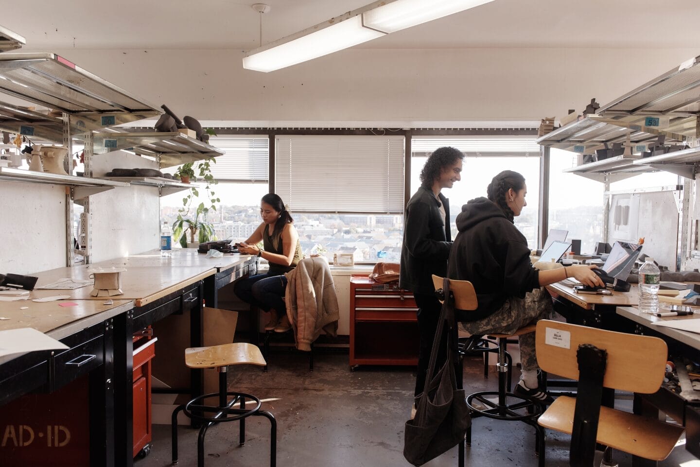 In an industrial design studio, three people work diligently. One stands while two are seated, engrossed in laptops and papers. Shelves brim with various items, and large windows offer city views, enhancing the creative atmosphere.