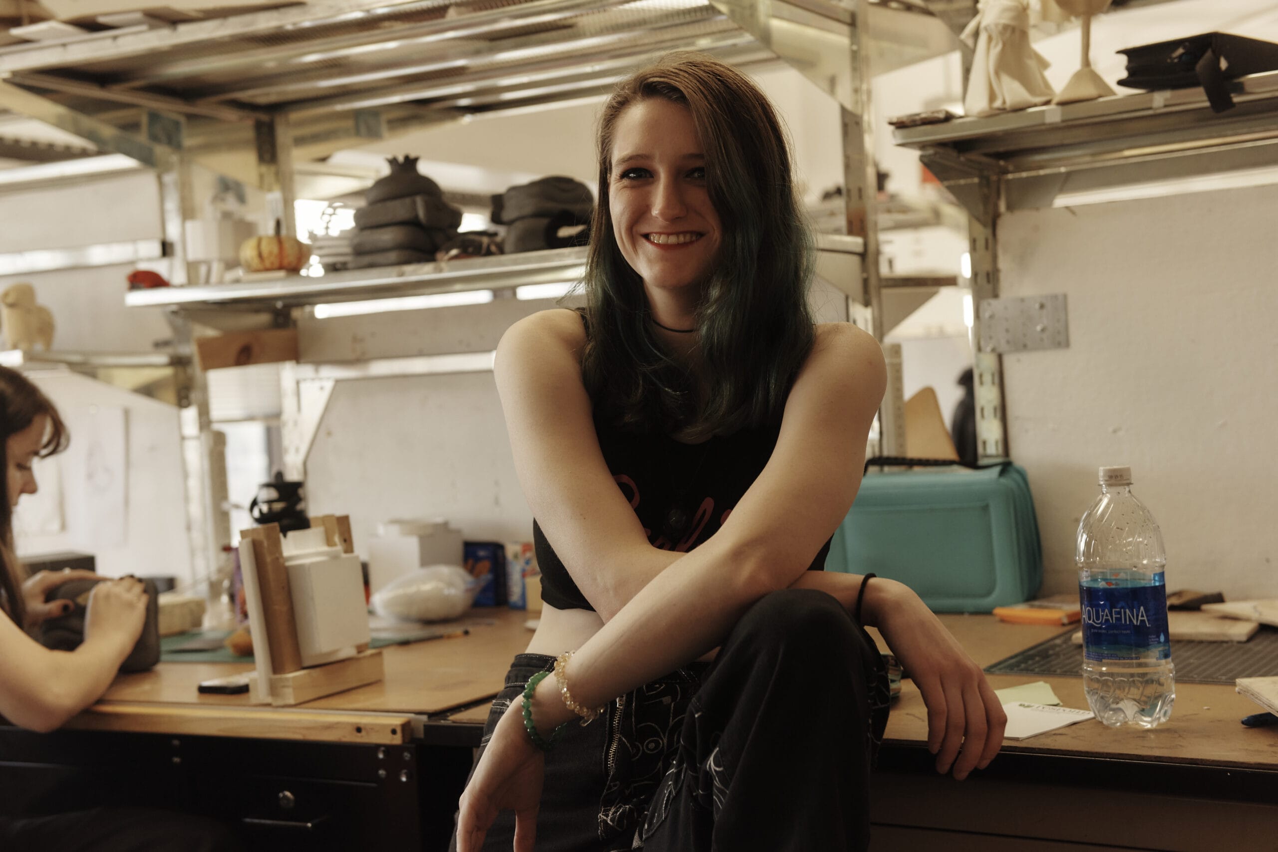 In a room reminiscent of a design studio, a person with long hair sits on a workbench surrounded by shelves. Smiling in their sleeveless top, they have crossed legs, and next to them is a bottle of water. In the background, another individual focuses at the desk—a snapshot of industrial design creativity in action.