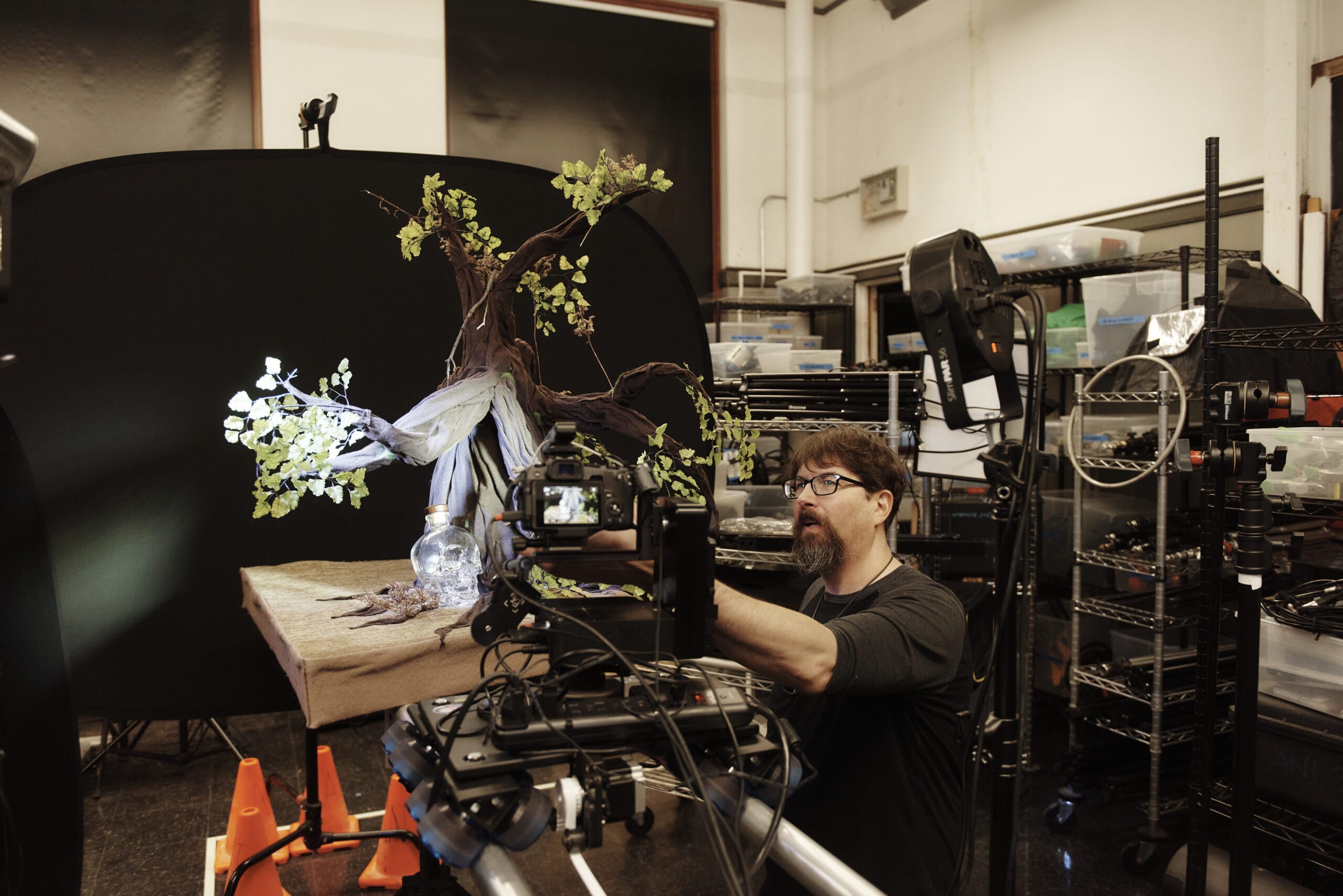 A person crouched in front of an animation camera with scenery on a table behind him.
