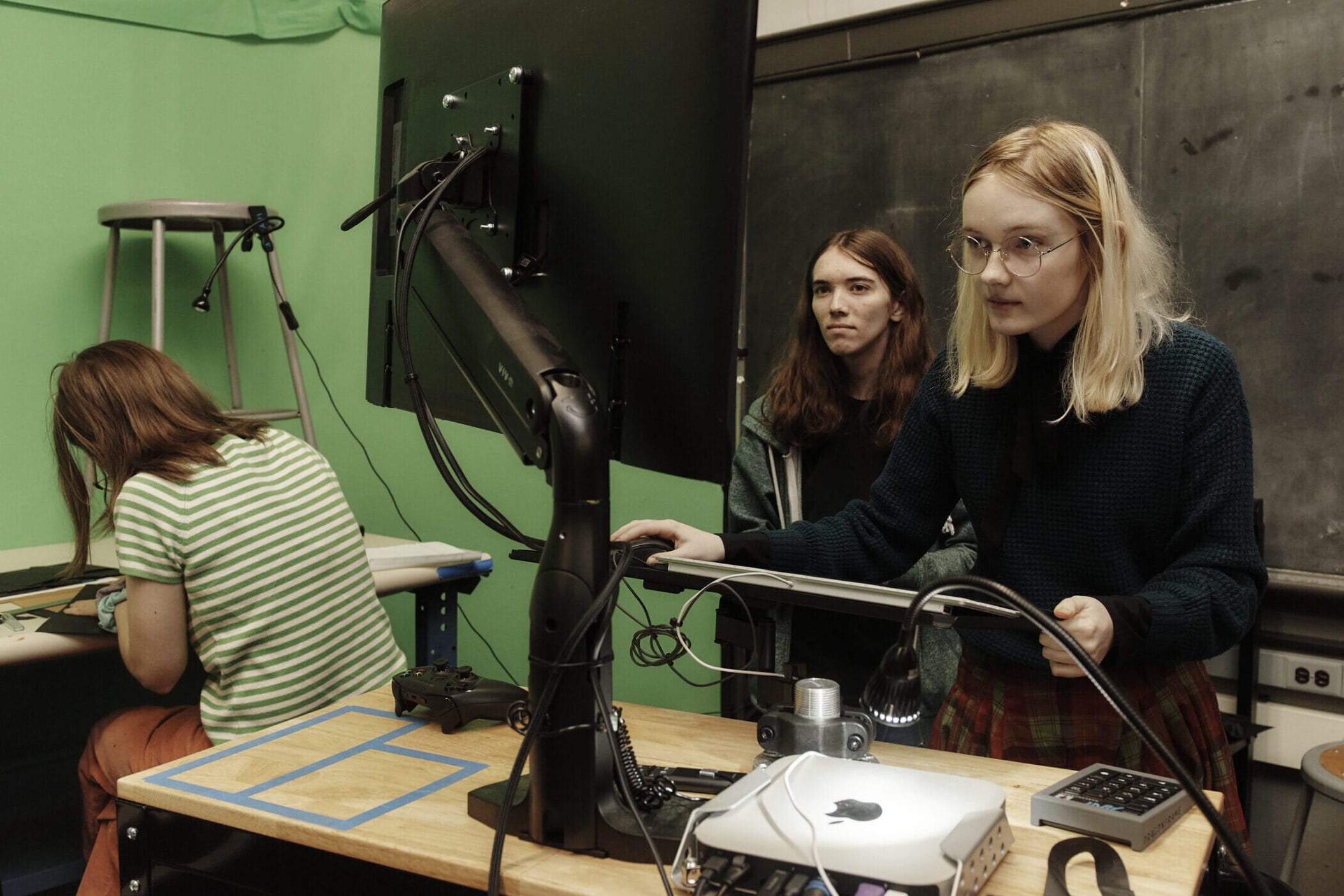 Three people in a room with a green screen. One person is seated at a table with electronics, another stands watching, and the third stands at a computer with monitors and wires. A ladder is in the background near a chalkboard.