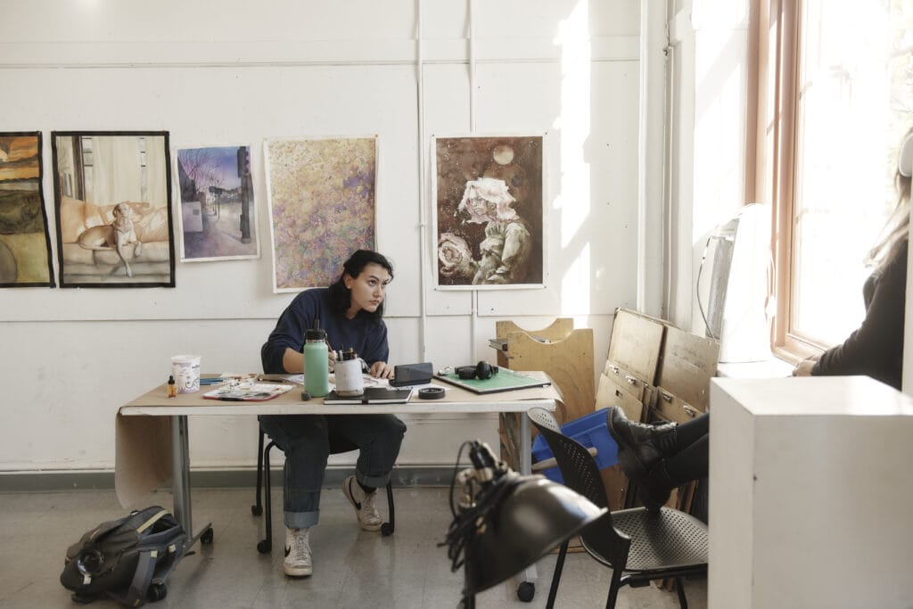 A student sitting a table, drawing, while looking up at a student posing in a window.