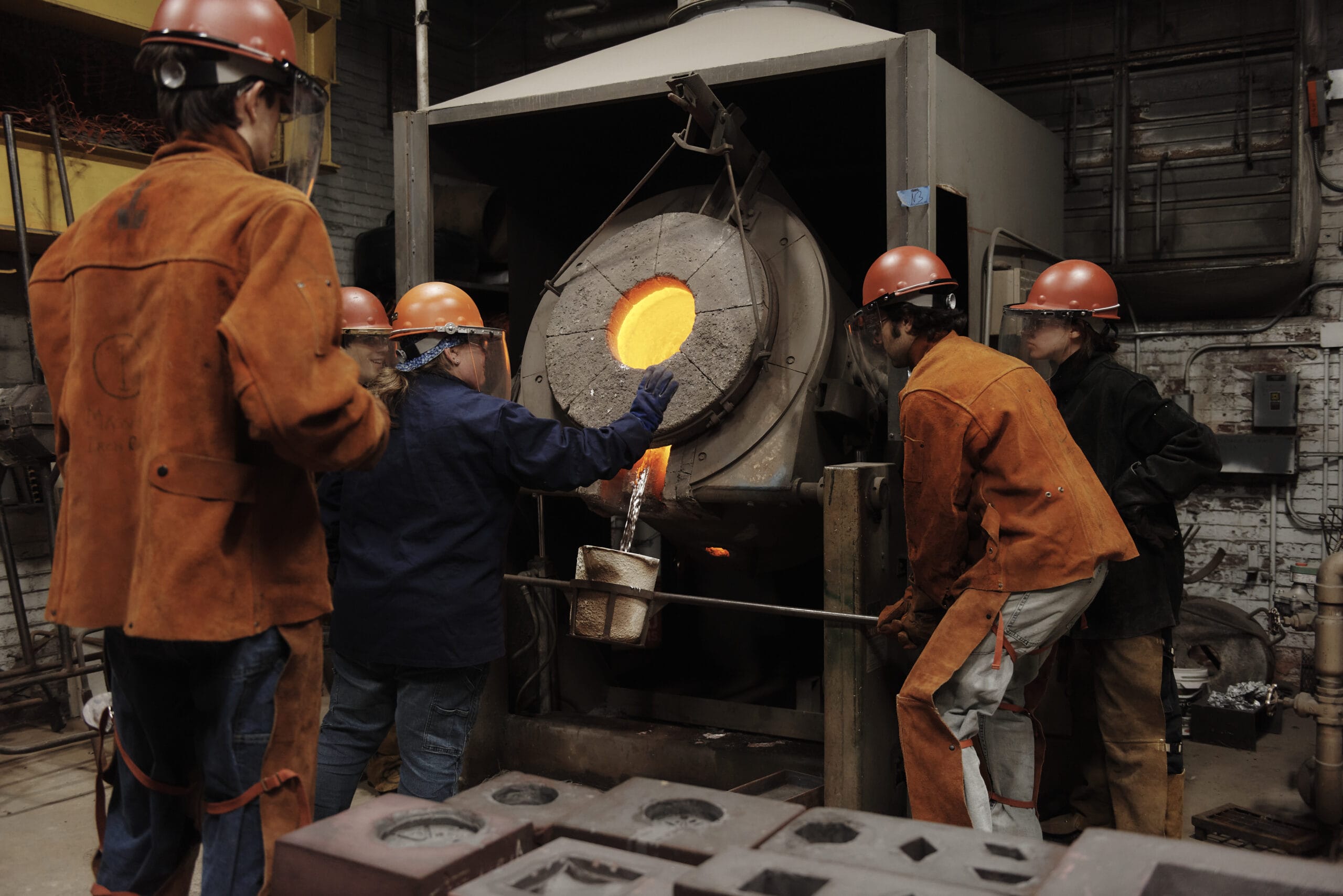 A group of workers wearing protective gear and helmets are gathered around a large, glowing furnace in an industrial setting. One person skillfully pours molten metal into a mold, shaping the beginnings of a sculpture, while others observe.