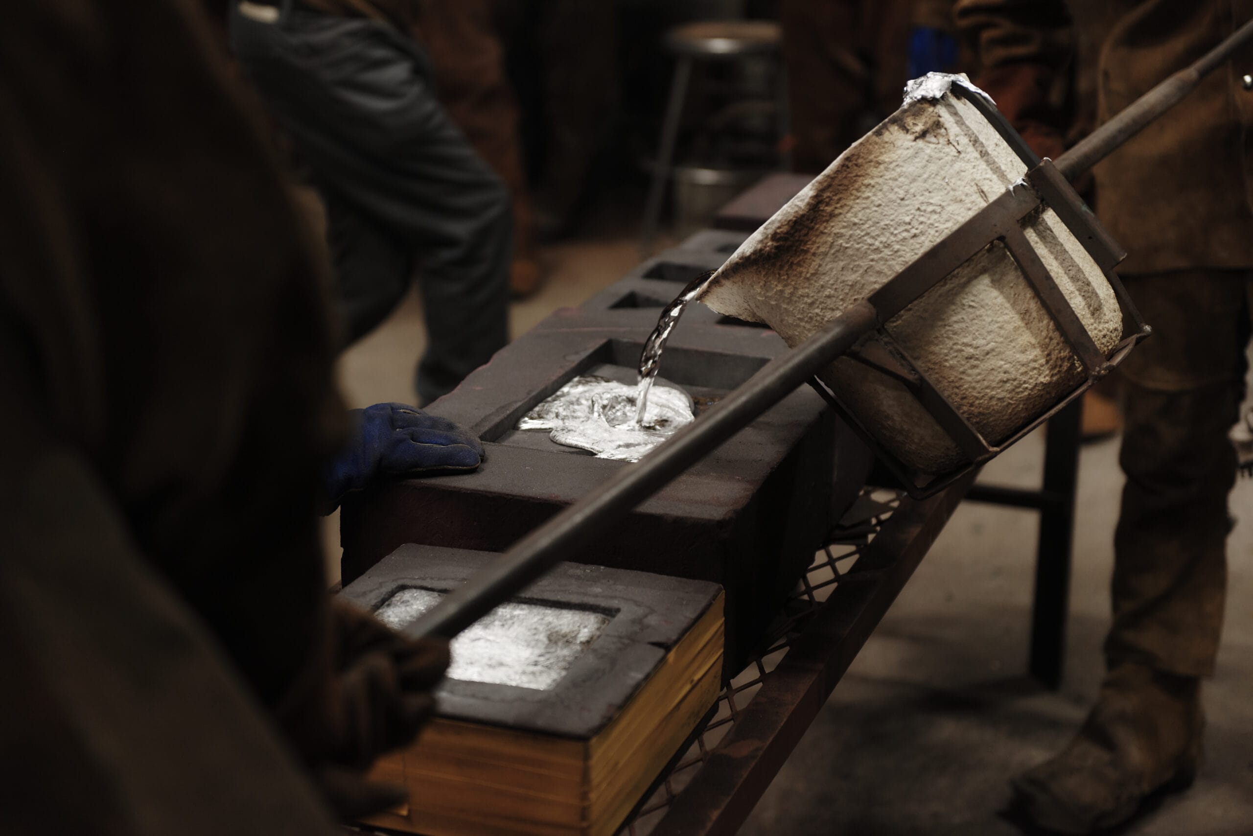 A worker pours molten metal into a mold using a large, handled crucible in an industrial sculpture workshop. The mold sits on a metal table, bordered by other molds. Workers clad in protective gear bustle in the background, utilizing shared resources to craft their creations.