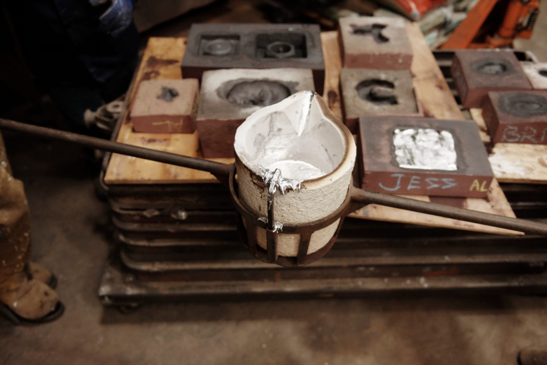 In an industrial environment, a metal casting setup with a ceramic crucible brimming with molten silver is supported by a sturdy metal frame. Surrounding it are various molds and casts labeled with names, like intriguing sculptures in the making—a testament to resourceful artistry.