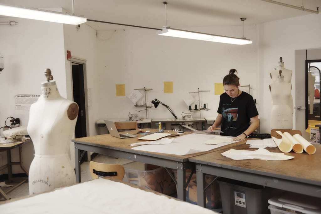 In a bustling fashion design studio, a person skillfully cuts fabric patterns on a large table, surrounded by sewing machines and mannequins. The well-lit room is alive with scattered paper rolls and crafting materials, echoing the creativity found in MassArt Fashion Design Studios.