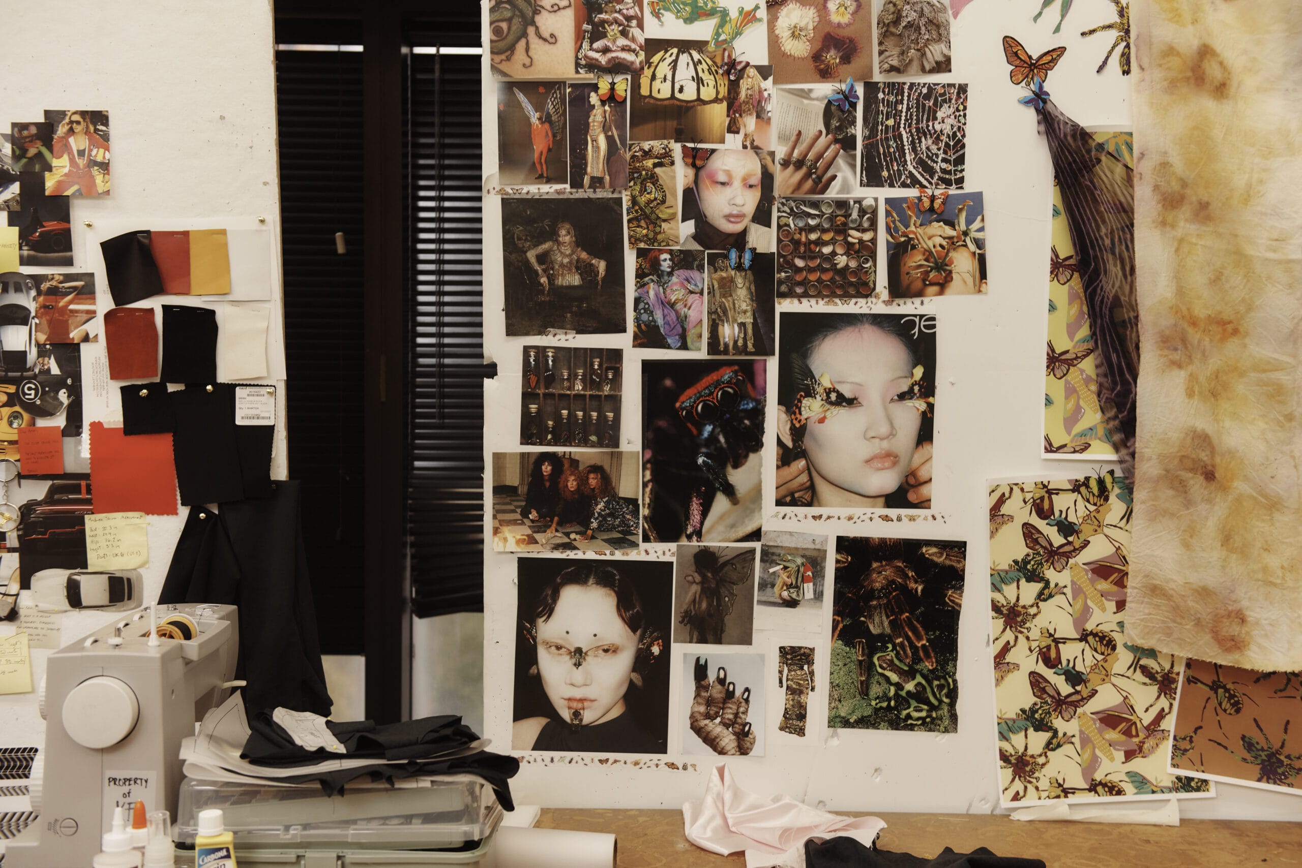 A creative fashion design studio with a wall adorned in fashion and art photos featuring models with unique makeup. MassArt Fashion Design Studios come alive with fabric swatches and designs, alongside a sewing machine and assorted materials on a vibrant table.