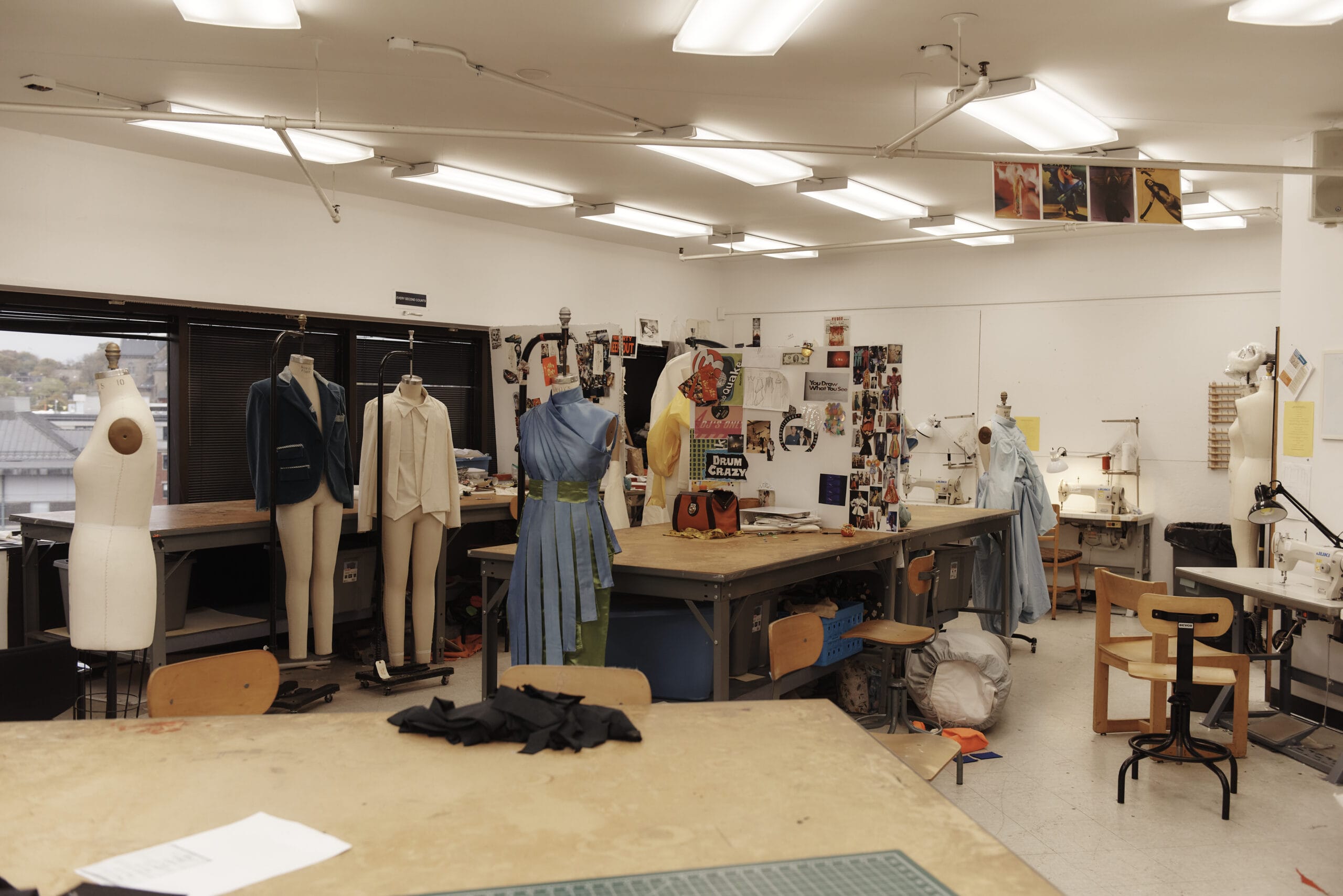 A spacious creative fashion design studio features multiple dress forms, sewing machines, and garments. The well-lit room is filled with tables covered in materials and bulletin boards adorned with fashion sketches. Large windows offer a captivating outside view, reminiscent of the inspiring MassArt Fashion Design Studios.
