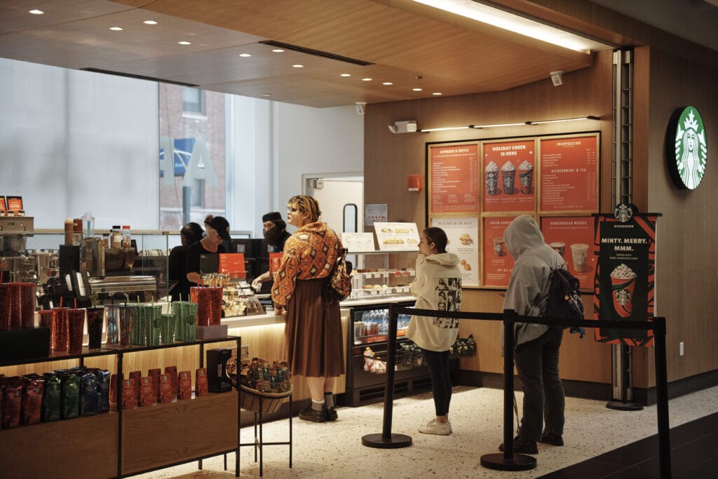 People stand in line inside a Starbucks, waiting to order. The cozy dining area features a large menu board, holiday-themed decorations, and a display with various colorful packaged items.