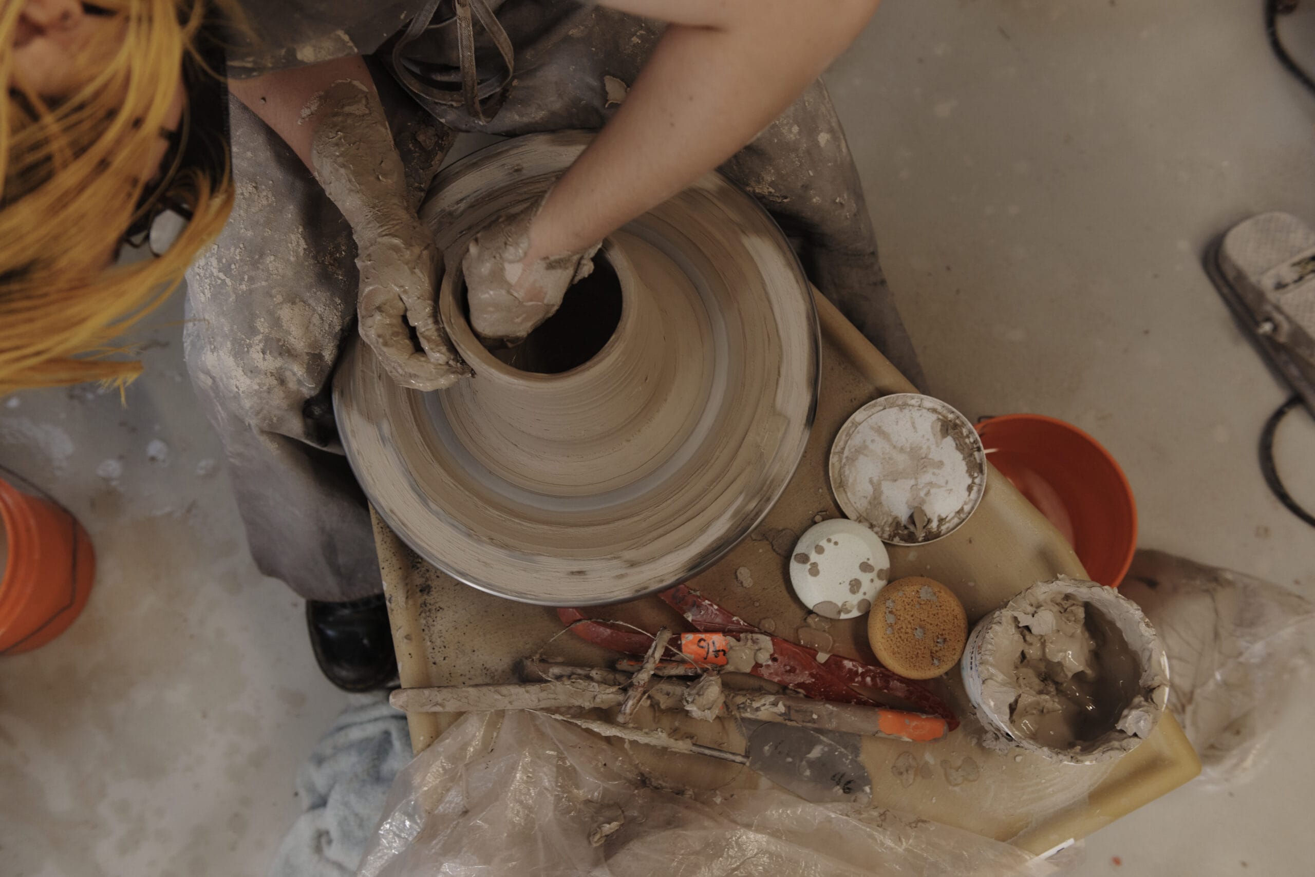A person wearing a brown apron and gloves shapes clay on a pottery wheel, embodying the spirit of MassArts creative education. Various pottery tools and containers with clay slip are arranged on a wooden tray nearby, while the floor is splattered with clay.