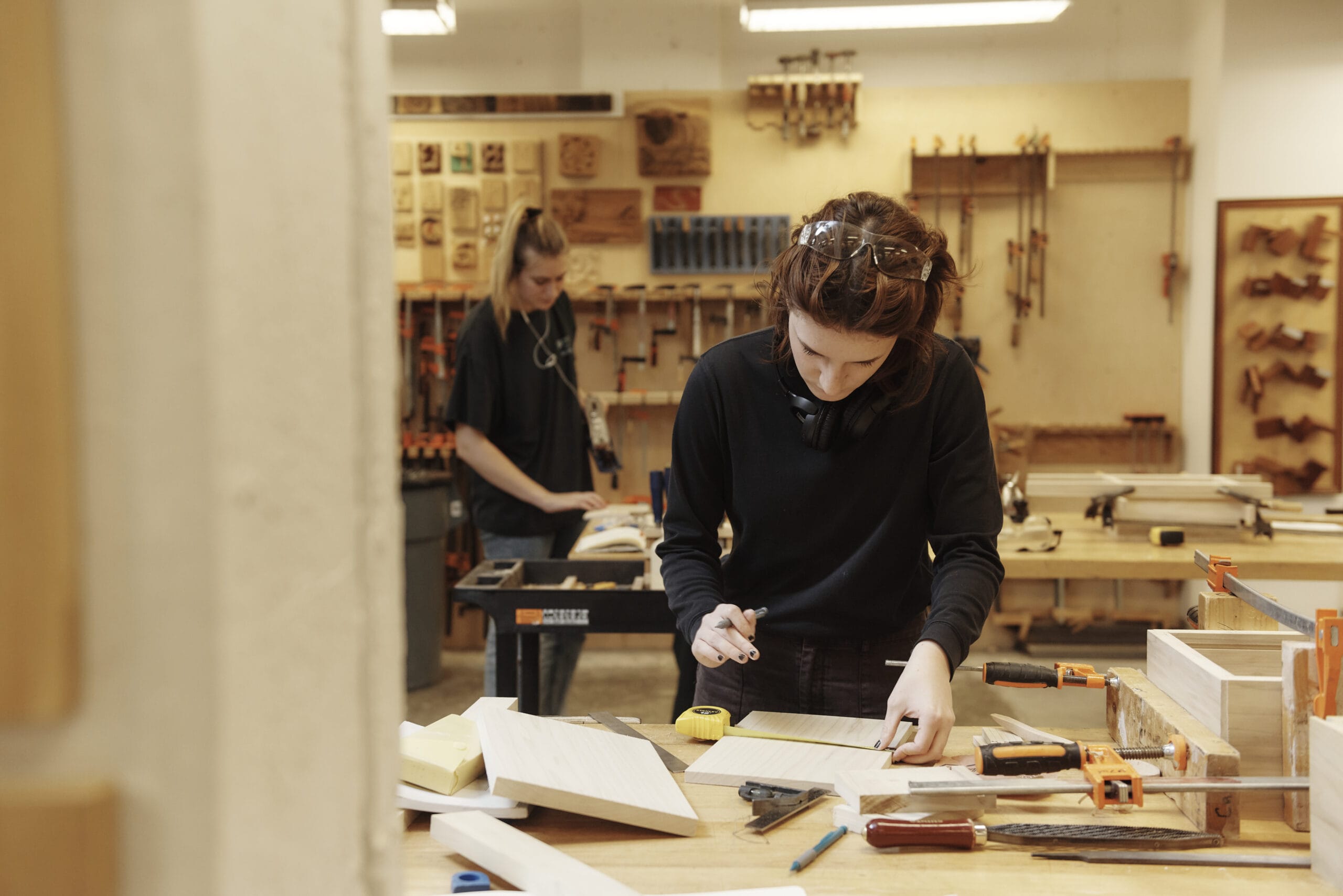 Two people immersed in crafting furniture in a woodworking shop. The person in the foreground meticulously measures wood amid a table cluttered with tools, while their partner, focused on design details in the background, creates harmony. Tools and wooden creations adorn the walls.
