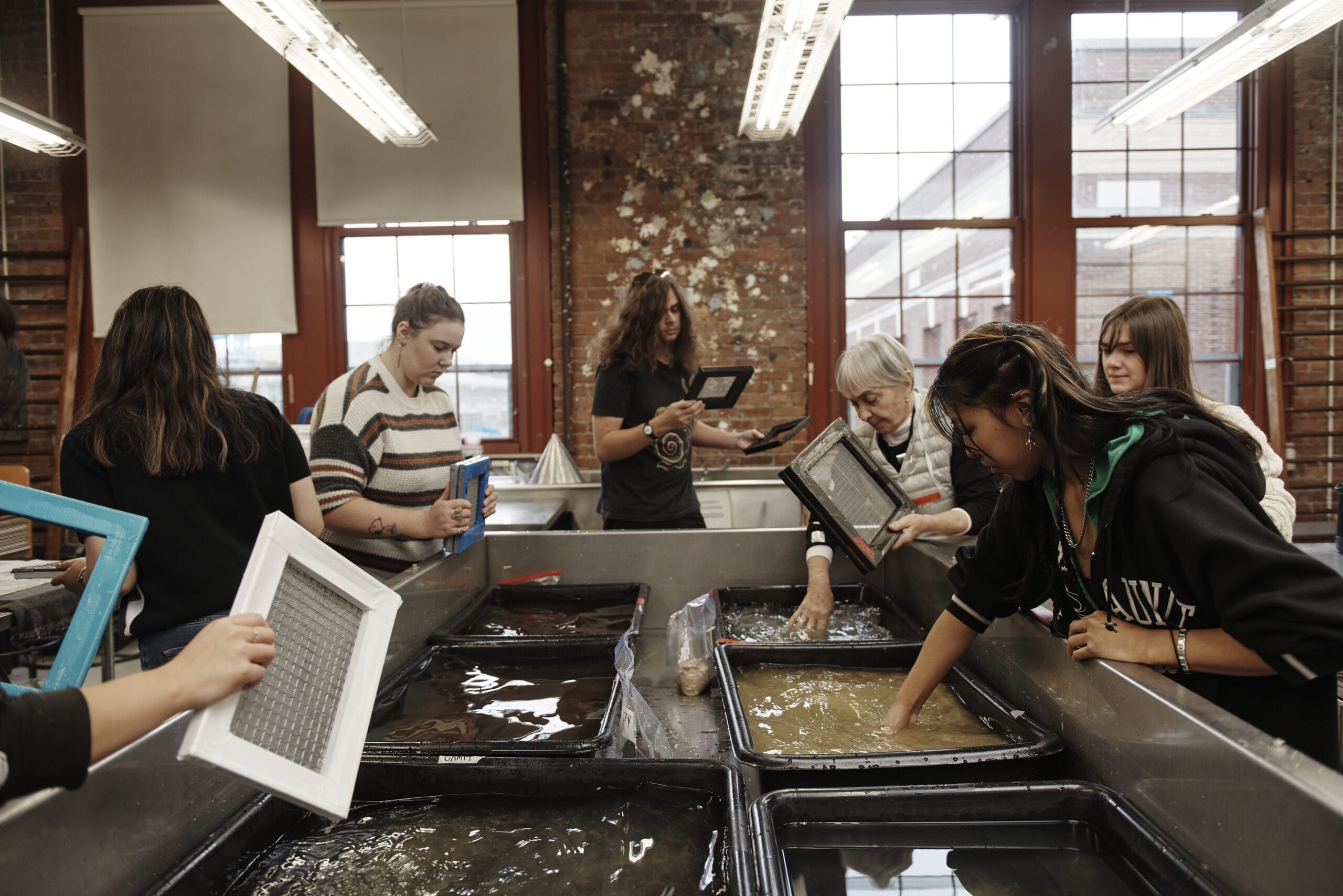 A group of people participates in a papermaking workshop. They are gathered around trays filled with liquid pulp, using screens to shape sheets of paper. The room has exposed brick walls and large windows.