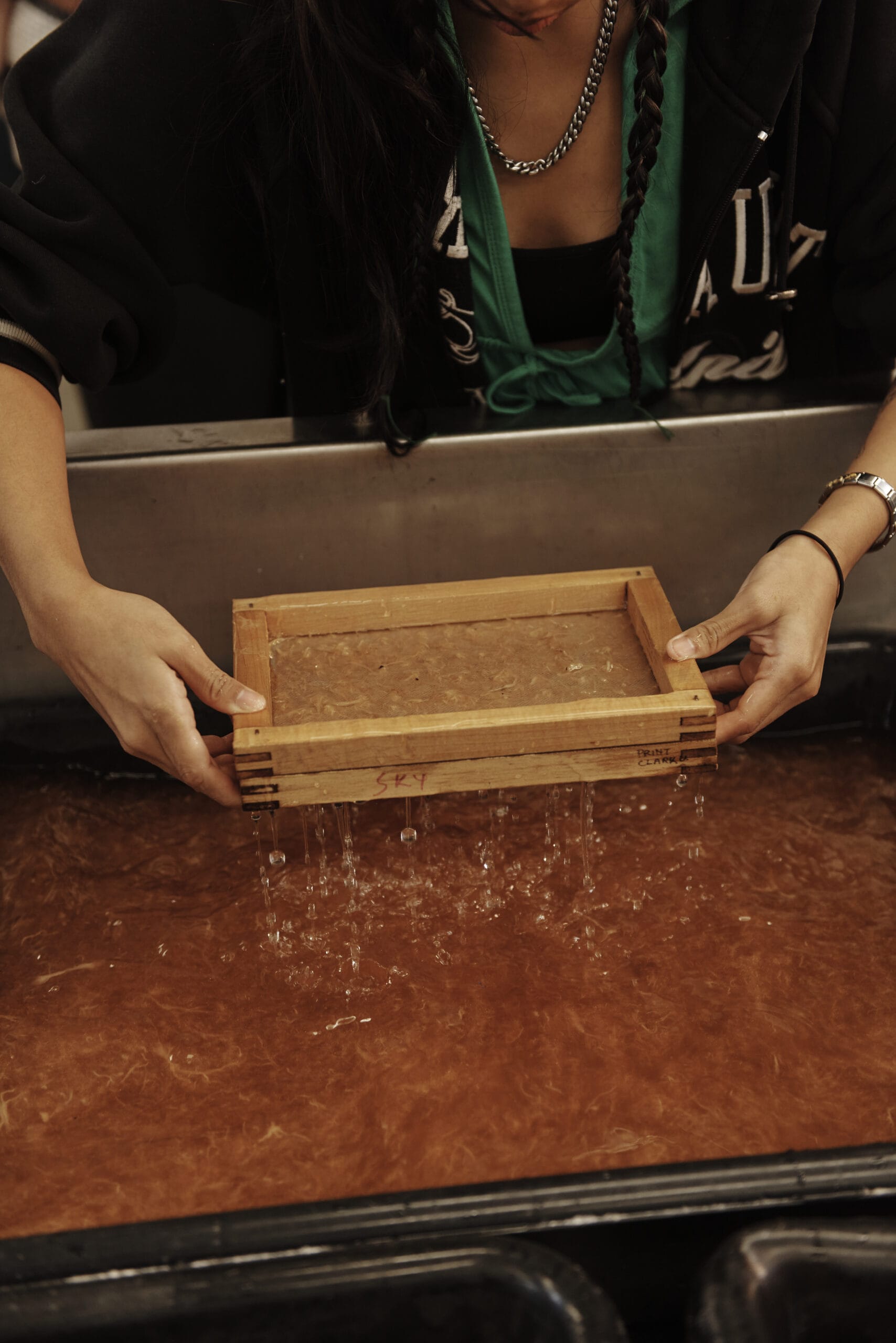 A person is making handmade paper using a wooden frame and mold, dipping it into a basin filled with water and pulp. The person is wearing a black hoodie, green shirt, and a chain necklace.