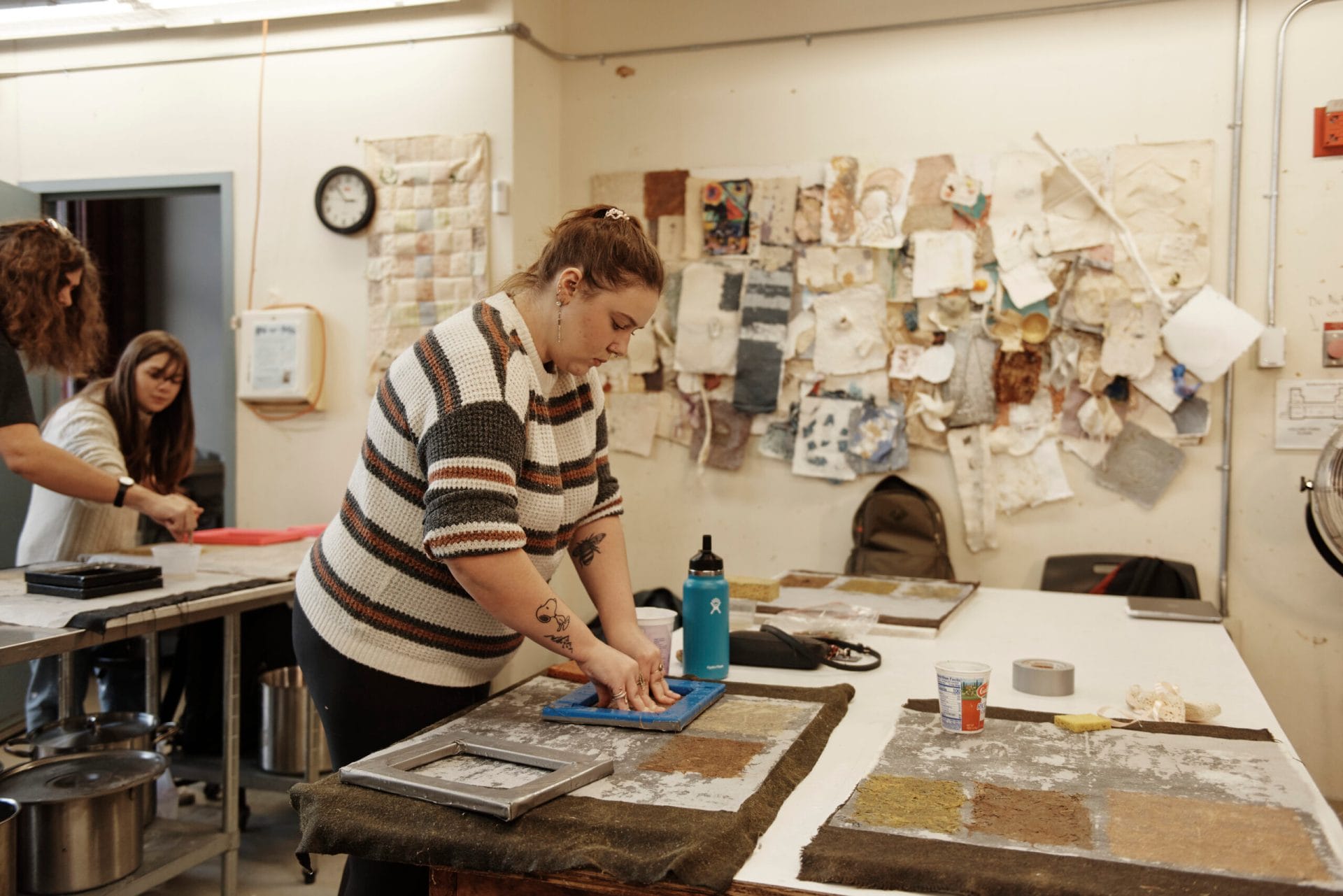 A woman in a striped sweater works on a project at a table in an art studio. Art supplies and materials are scattered around. Another woman works in the background. The walls display a variety of textured art pieces.