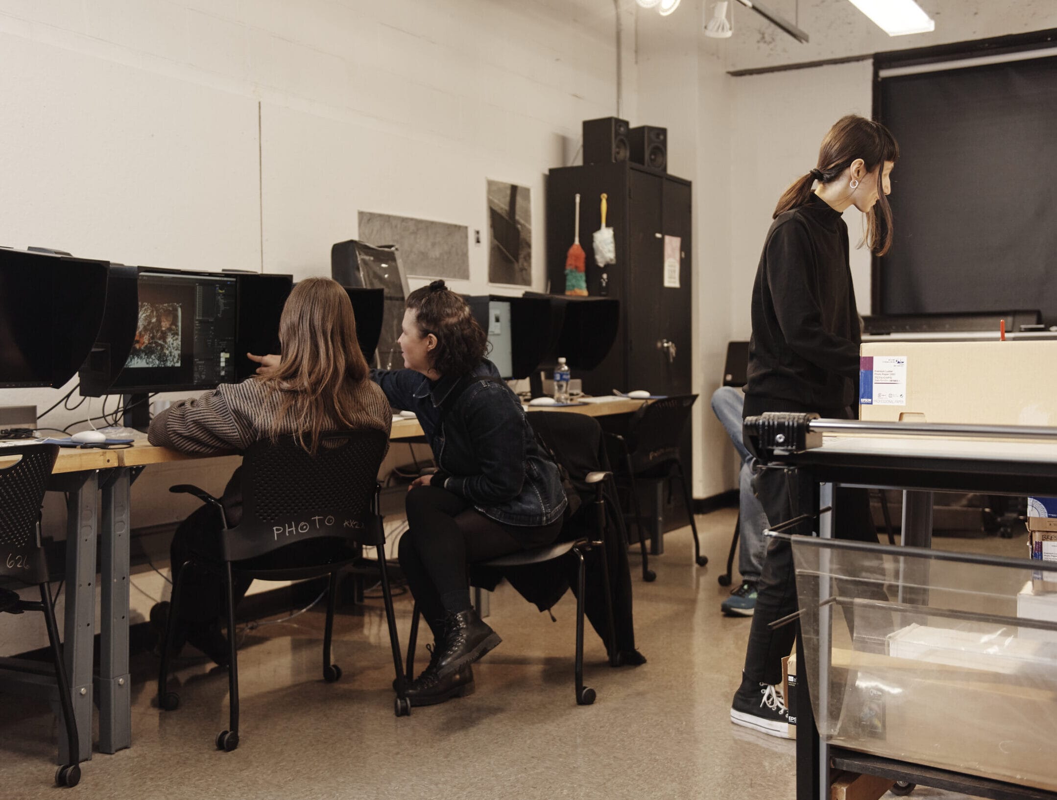 Two students sit in front of computer monitors in a digital photography lab while one stands at a table.