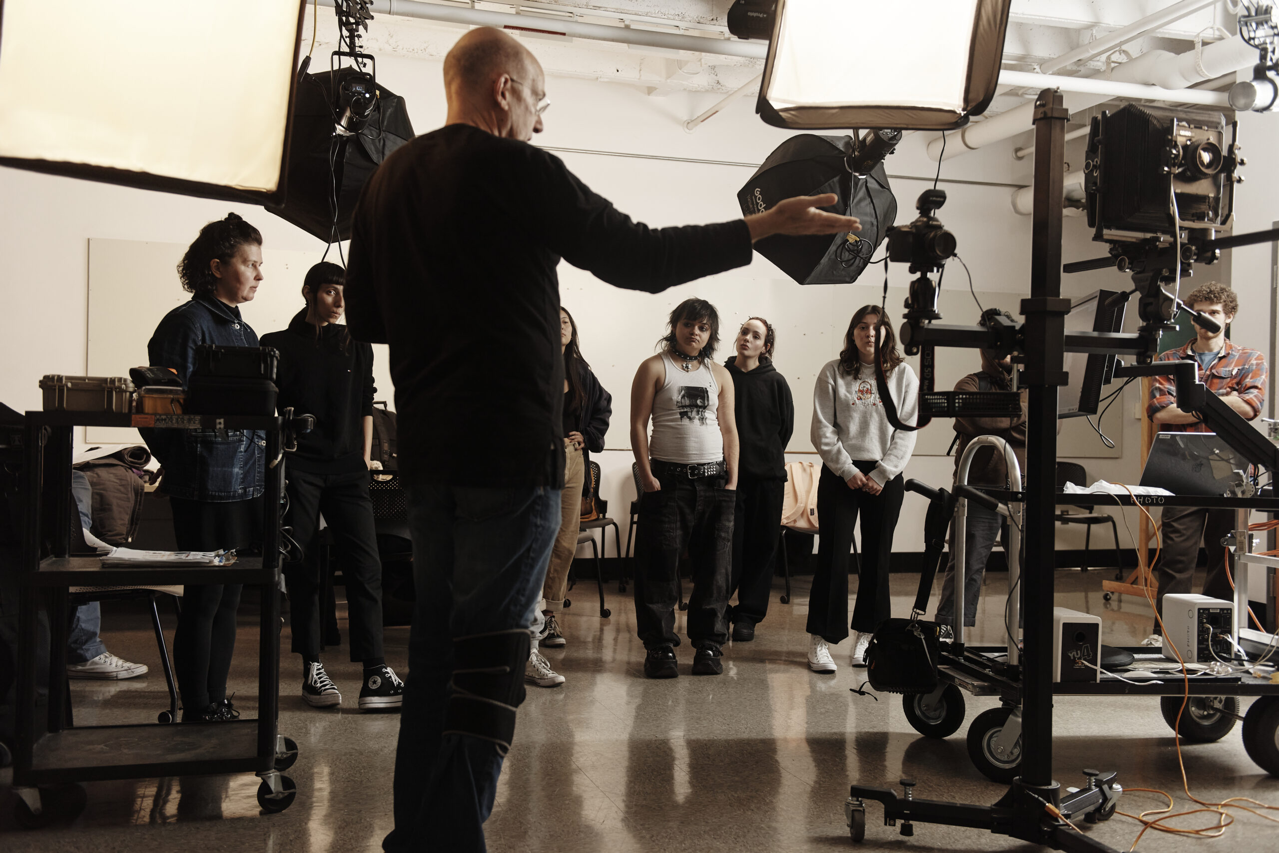 In a studio setting bathed in professional lighting, a lively individual gestures animatedly, instructing the group amidst an array of photography gear. Behind them, the digital lab hums with creativity, adding a modern touch to the art form.