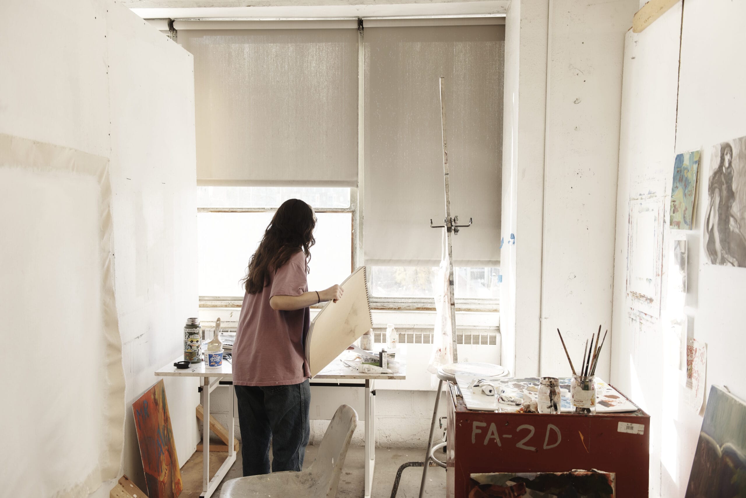 A person stands in a bright art studio, holding a canvas, the essence of their BFA journey in Fine Arts. The room is filled with supplies, paintings, and brushes. Large windows with blinds allow soft natural light to enter this inspiring space for painting masterpieces.