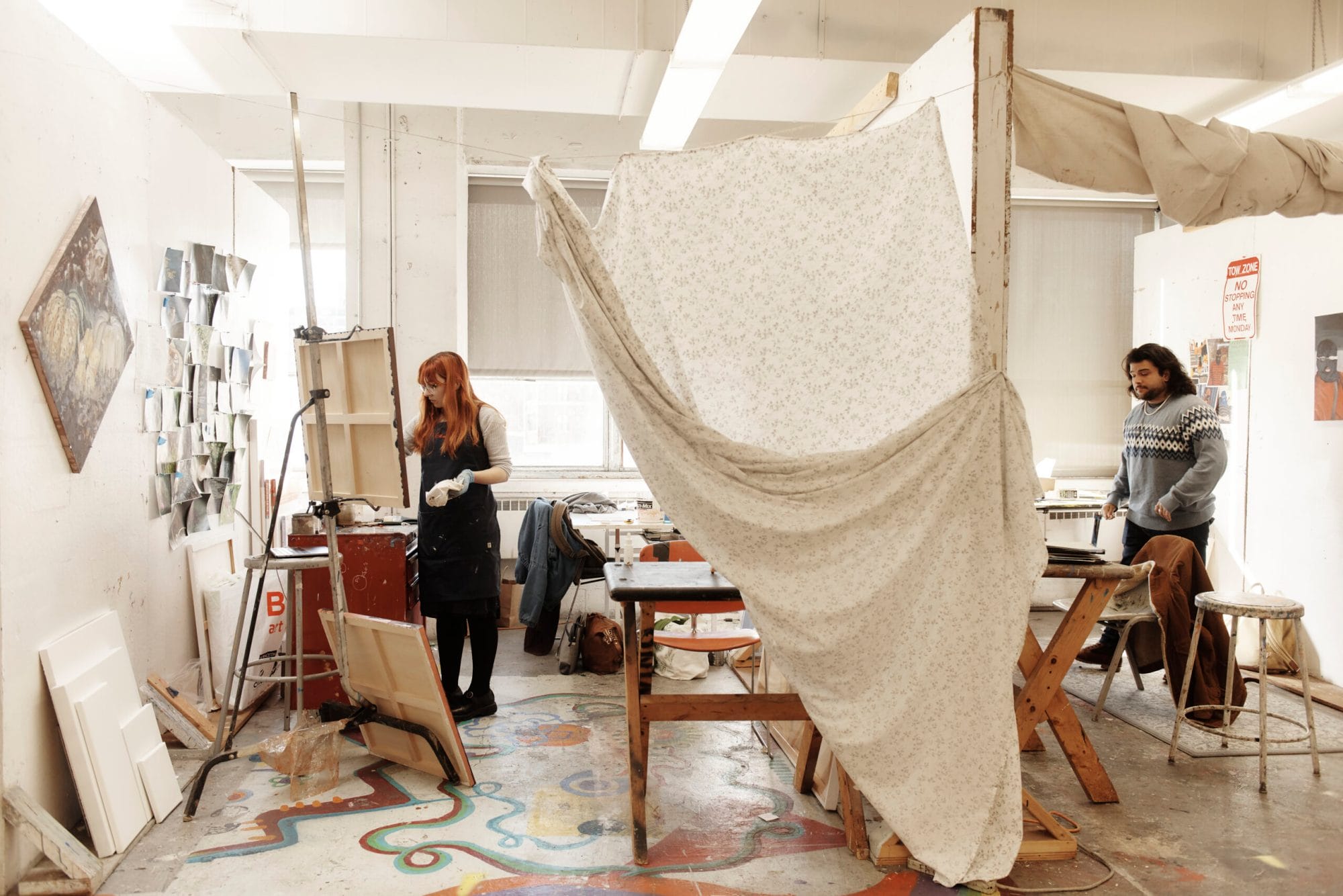 A curtain hangs down through the middle of the photo dividing two painting studios; a woman stands on the left and a man on the right in their studios.