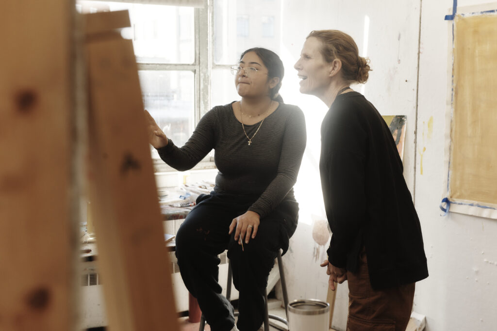 A teacher leans in to give advice to a student on a painting.